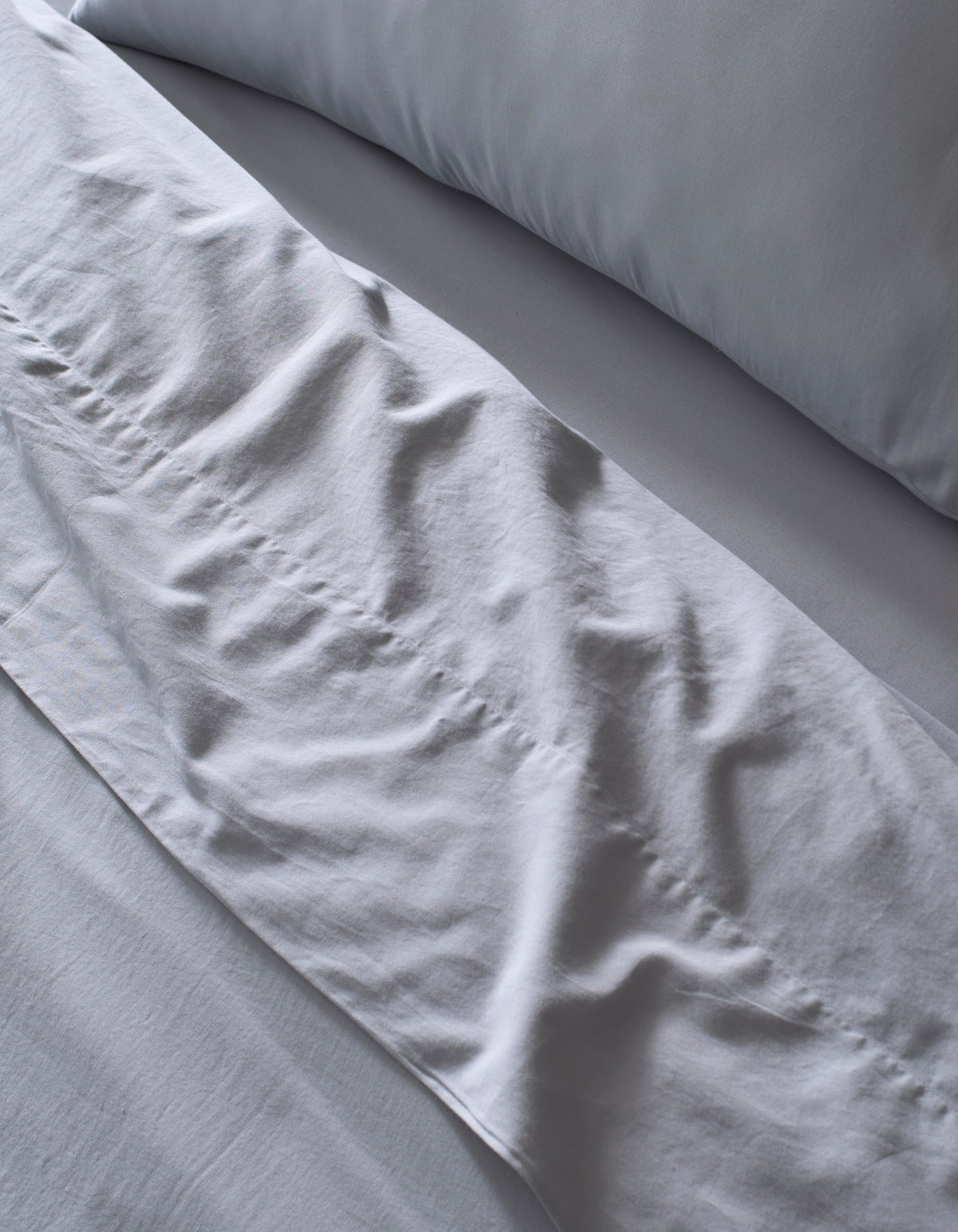 Close-up of a neatly made bed featuring the Cozy Earth Soft-Wash Cotton Bedding Core Bundle in light gray, with softly wrinkled sheets and pillowcase hinting at recent use or adjustment.|Color:Shore