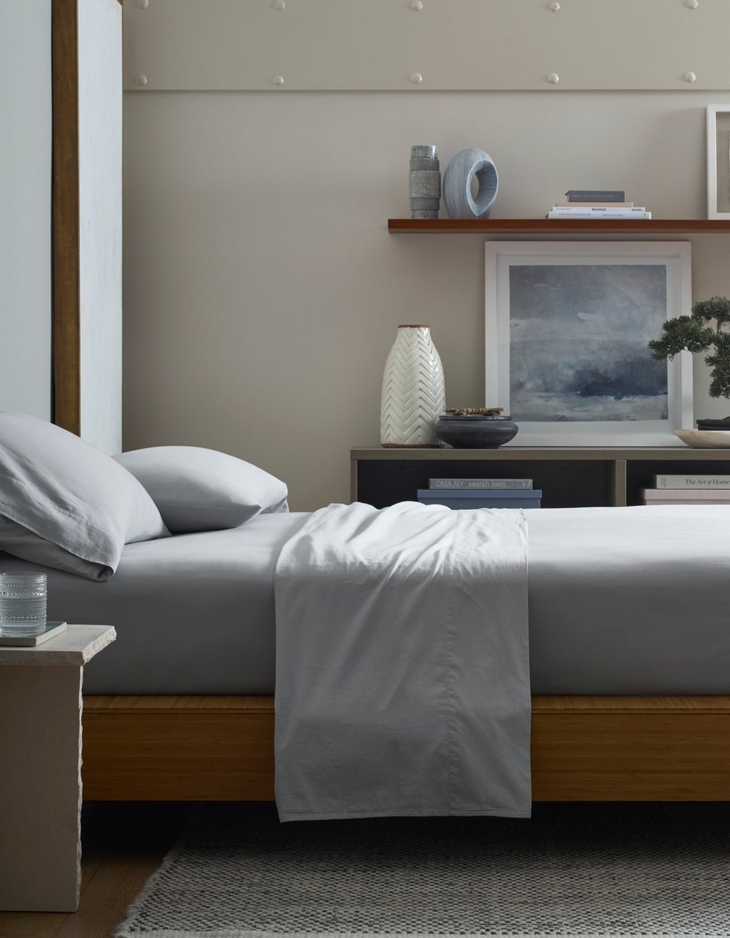 A neatly made bed with Cozy Earth Soft-Wash Cotton Flat Sheets sits by a nightstand holding a glass of water. Shelves behind display books, vases, and framed art, creating a serene and modern bedroom atmosphere.|Color:Shore