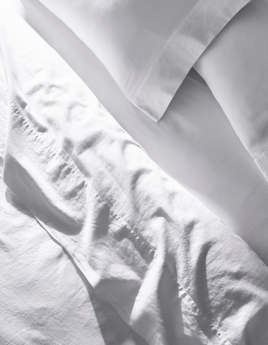 The Soft-Wash Cotton Sheet Set by Cozy Earth features slightly wrinkled white bed sheets and pillowcases neatly arranged on a bed, giving a clean, bright look in natural light.|Color:White