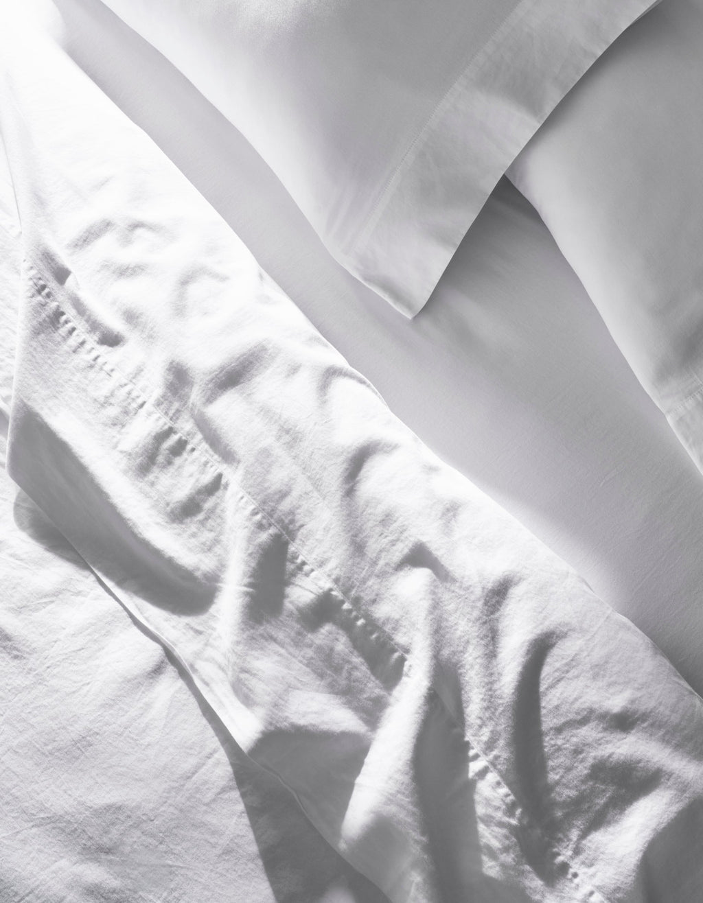 A close-up of the Cozy Earth Soft-Wash Cotton Bedding Core Bundle shows slightly wrinkled white sheets and pillowcases, with soft natural light casting gentle shadows and highlights on the fabric.|Color:White
