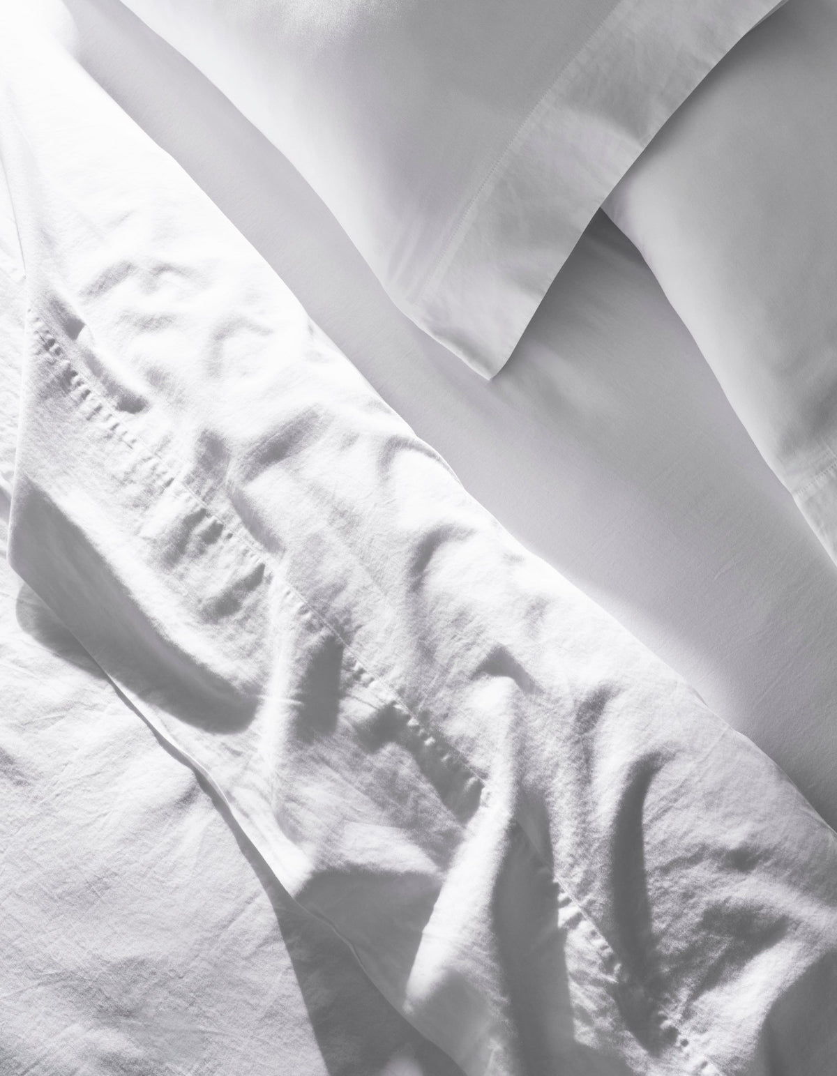 A close-up of the Cozy Earth Soft-Wash Cotton Bedding Core Bundle shows slightly wrinkled white sheets and pillowcases, with soft natural light casting gentle shadows and highlights on the fabric.|Color:White