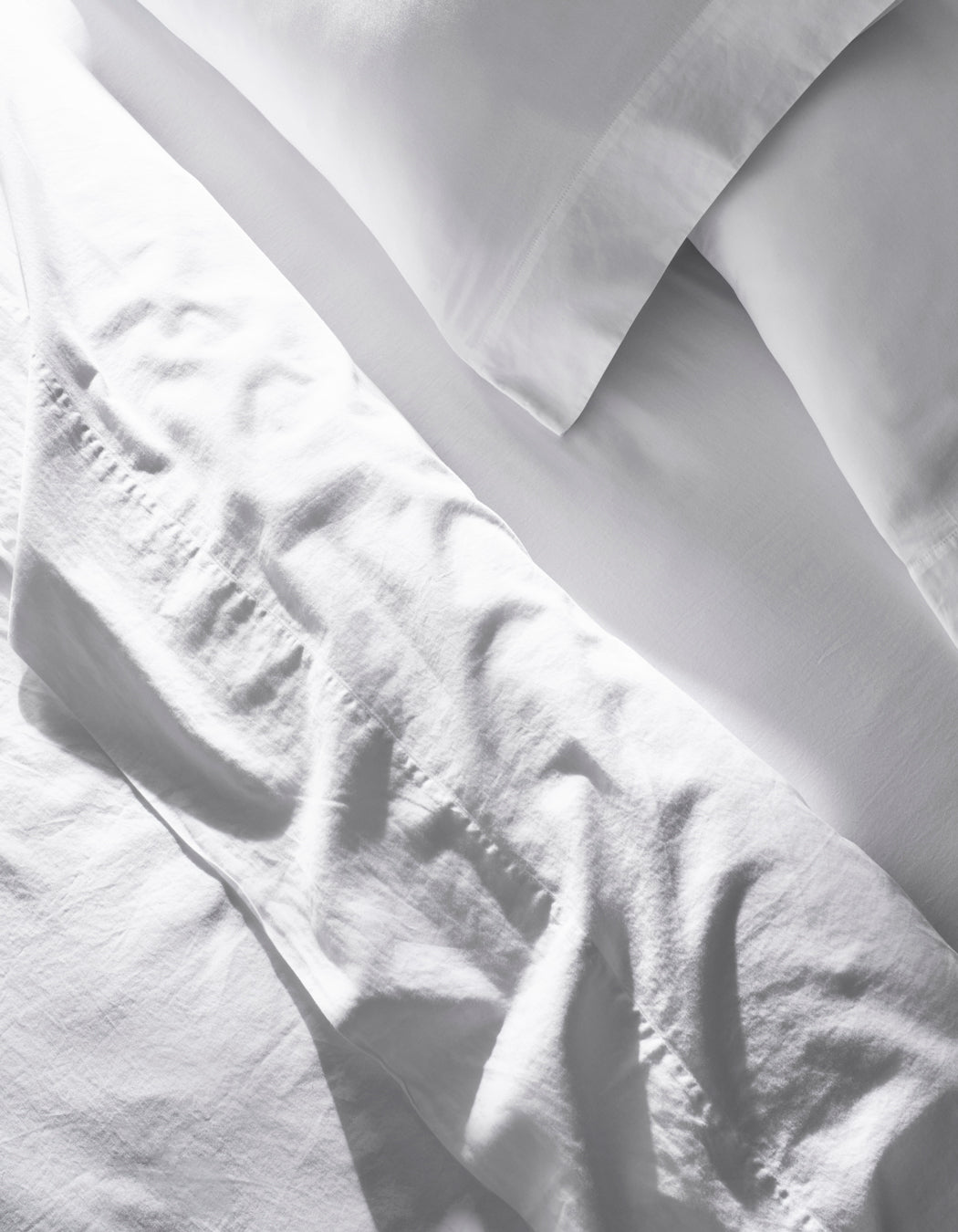 The Soft-Wash Cotton Sheet Set by Cozy Earth features slightly wrinkled white bed sheets and pillowcases neatly arranged on a bed, giving a clean, bright look in natural light.|Color:White
