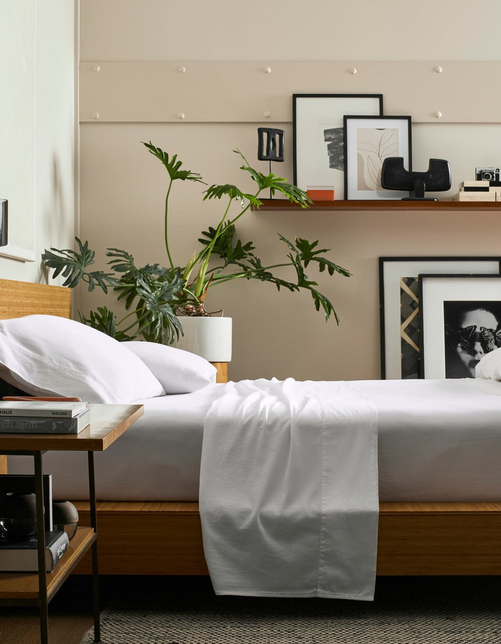 A modern bedroom features a wooden bed dressed in the Cozy Earth Soft-Wash Cotton Sheet Set, a bedside table with books, a large leafy plant in a white pot, and a shelf with framed art and decor against a beige wall.|Color:White