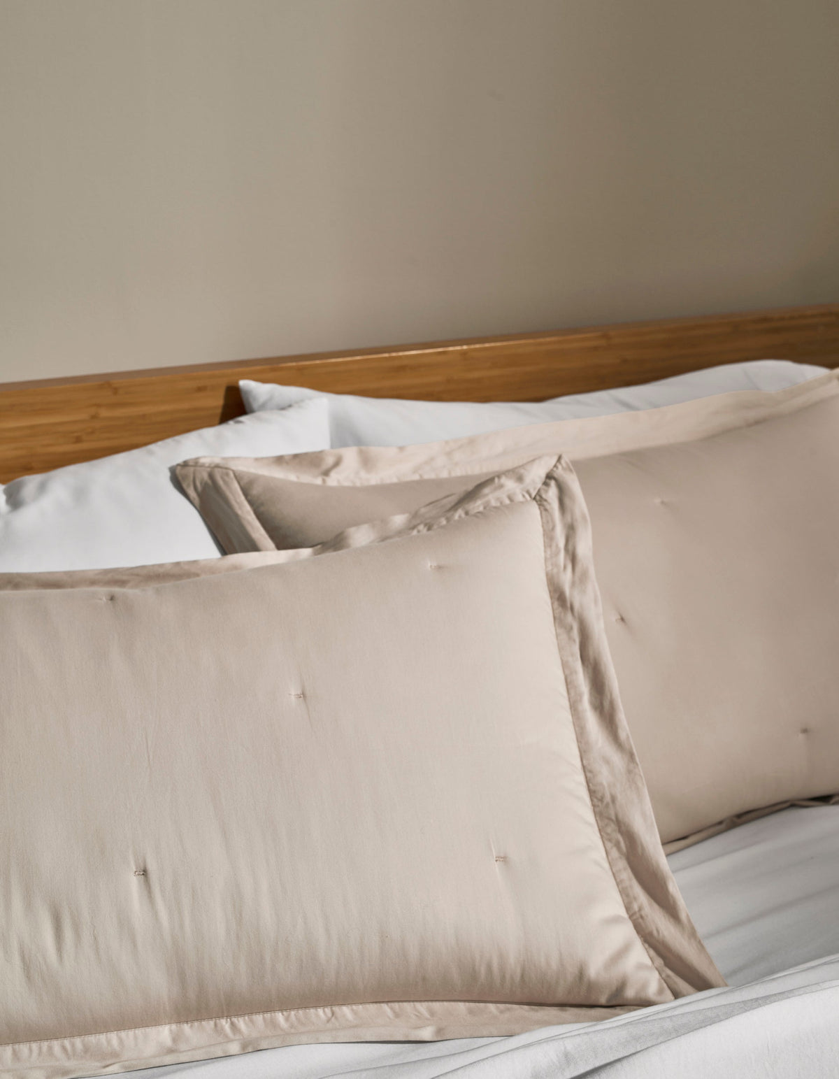 Two Cozy Earth Soft-Wash Cotton Quilted Shams in cream rest on a neatly made bed with white sheets and a wooden headboard against a beige wall.|Color:Driftwood