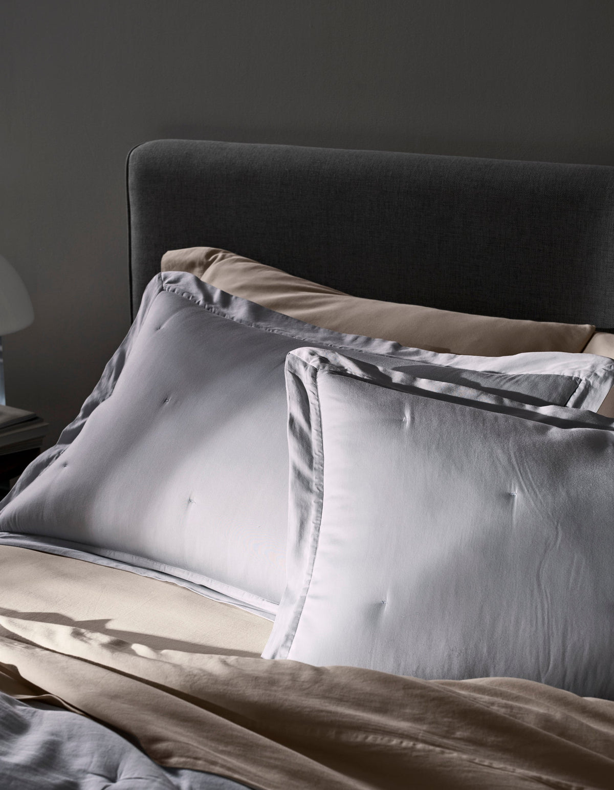 A close-up of an unmade bed features two Cozy Earth Soft-Wash Cotton Quilted Shams atop beige sheets and a dark gray upholstered headboard. Soft daylight highlights the calm, cozy atmosphere.|Color:Shore