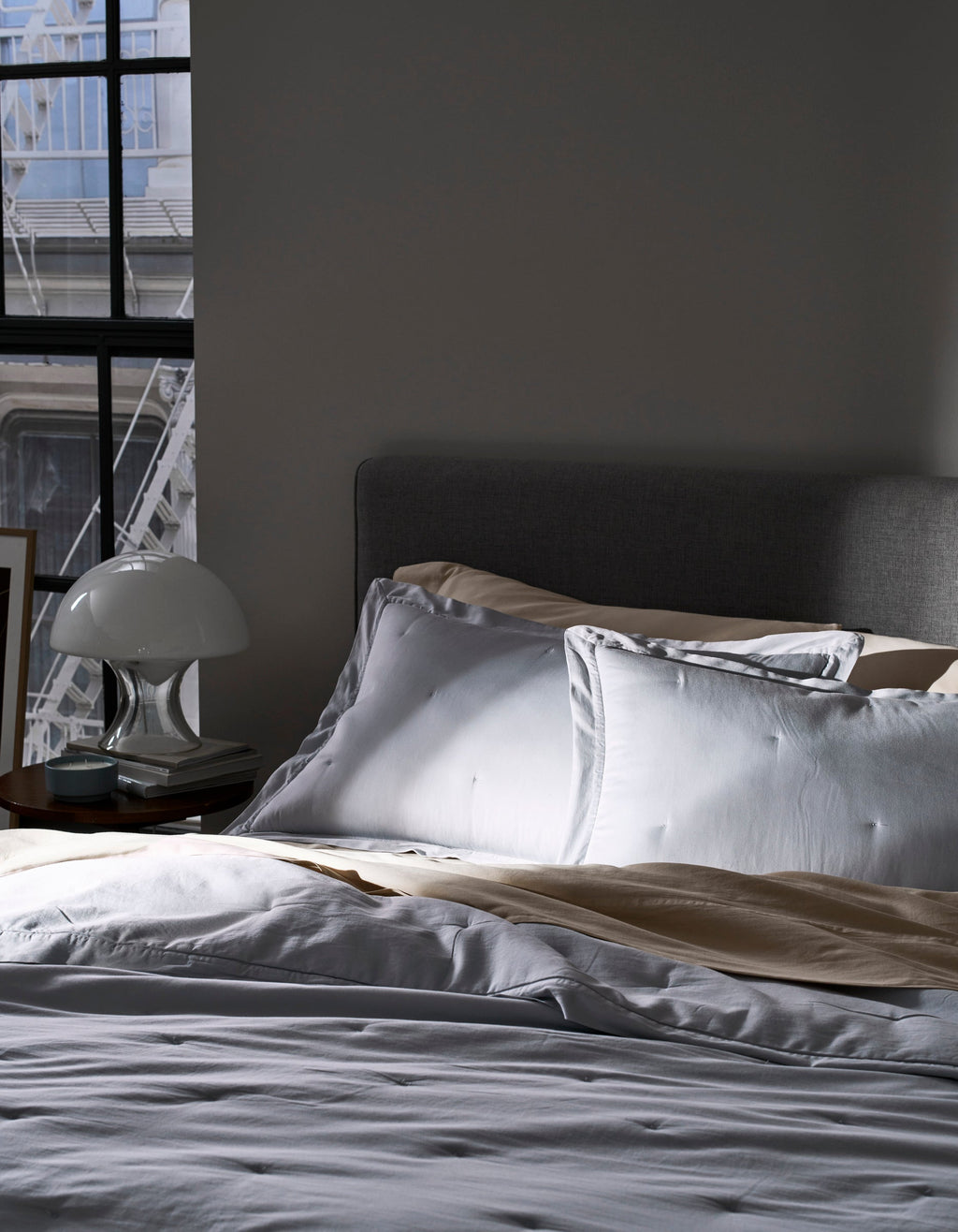 A bed with Cozy Earth Soft-Wash Cotton Quilted Shams in white and beige sits by a sunlit window. A bedside table holds a white lamp and books, while part of a gray upholstered headboard is seen.|Color:Shore