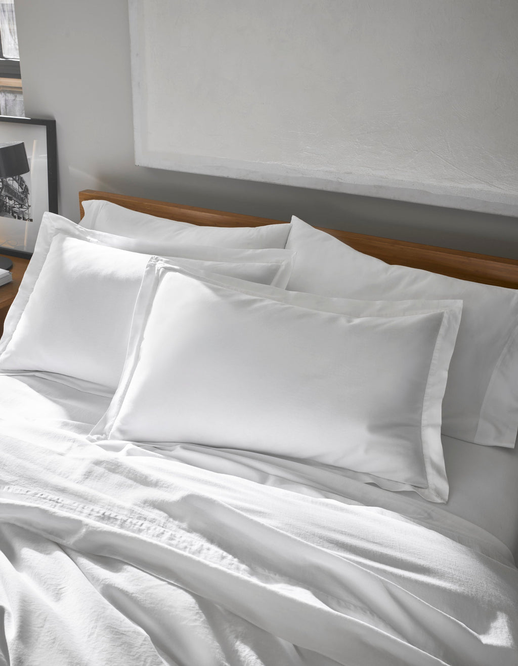 A neatly made bed features Cozy Earth's Soft-Wash Cotton Shams, crisp and white, paired with pillowcases against a wooden headboard in a softly lit, minimalist bedroom with a framed picture on the side table.|Color:White