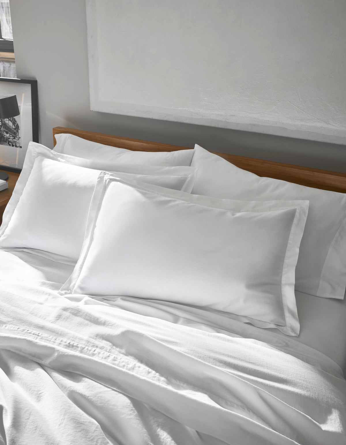 A neatly made bed features Cozy Earth's Soft-Wash Cotton Shams, crisp and white, paired with pillowcases against a wooden headboard in a softly lit, minimalist bedroom with a framed picture on the side table.|Color:White
