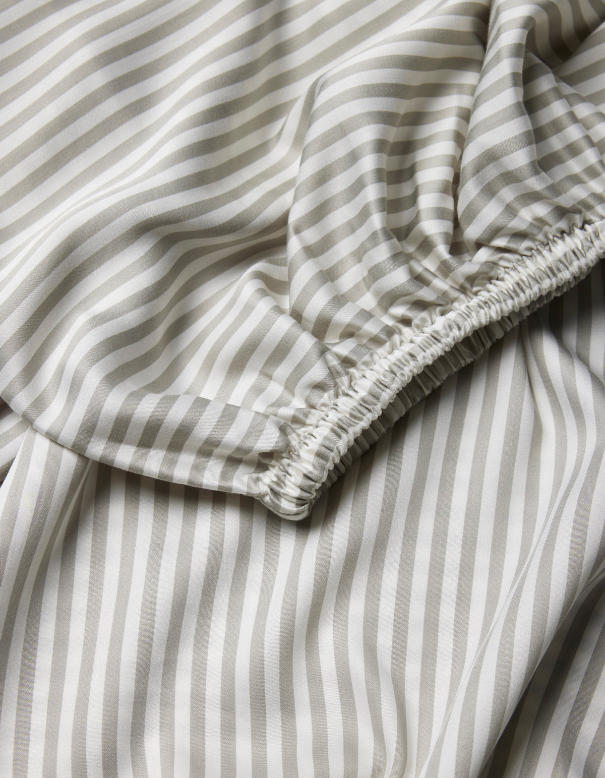 A close-up of Cozy Earth's Bamboo Fitted Sheet, showcasing light fabric with beige and white vertical stripes and a gathered, elasticized seam.|Color:Classic Stripe in Laurel