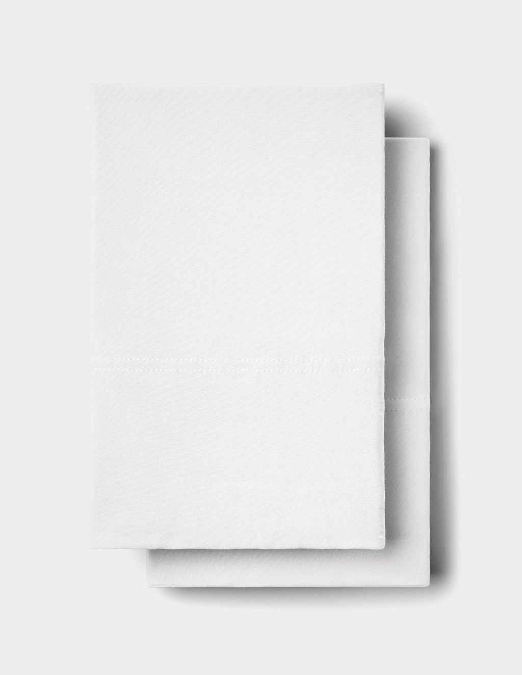 Two neatly folded Bamboo Jersey Pillowcases by Cozy Earth are stacked on a plain light gray surface, with one slightly overlapping the other. The pillowcases feature a subtle textured pattern and a visible horizontal crease line.|Color:White
