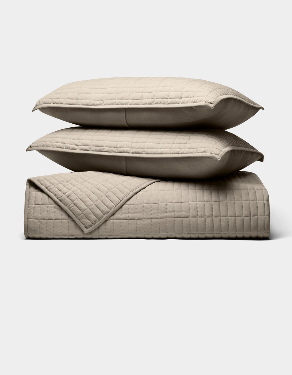The Cozy Earth Bamboo Jersey Quilt Bundle, shown against a white background, features two beige quilted pillows stacked on a matching blanket with a simple square-stitched design.|Color:Driftwood