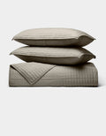 Two beige Cozy Earth Bamboo Jersey Quilt Bundle pillows are stacked atop a matching quilted blanket, all neatly arranged against a plain light background.|Color:Stone