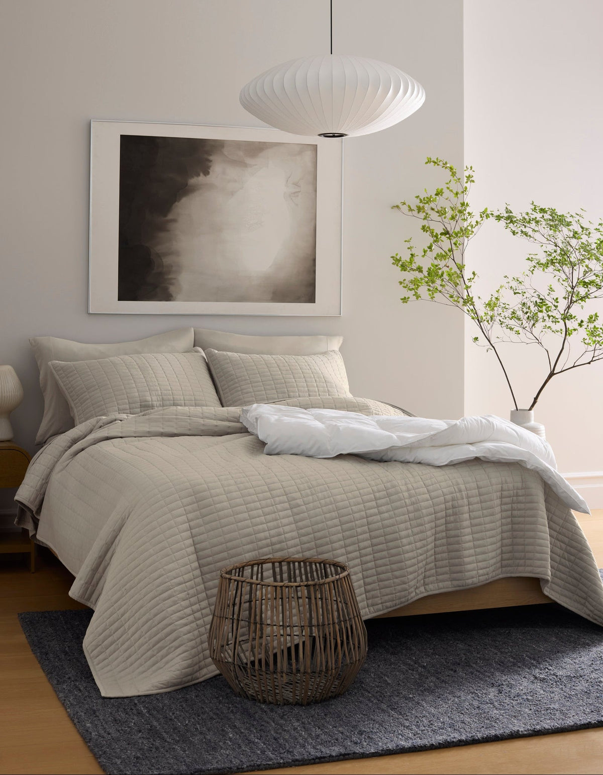 A modern bedroom featuring the Cozy Earth Bamboo Jersey Quilt in beige, a dark gray rug, a woven basket at the bed's foot, a minimalist pendant lamp, abstract wall art, and a small potted tree in the corner.|Color:Driftwood