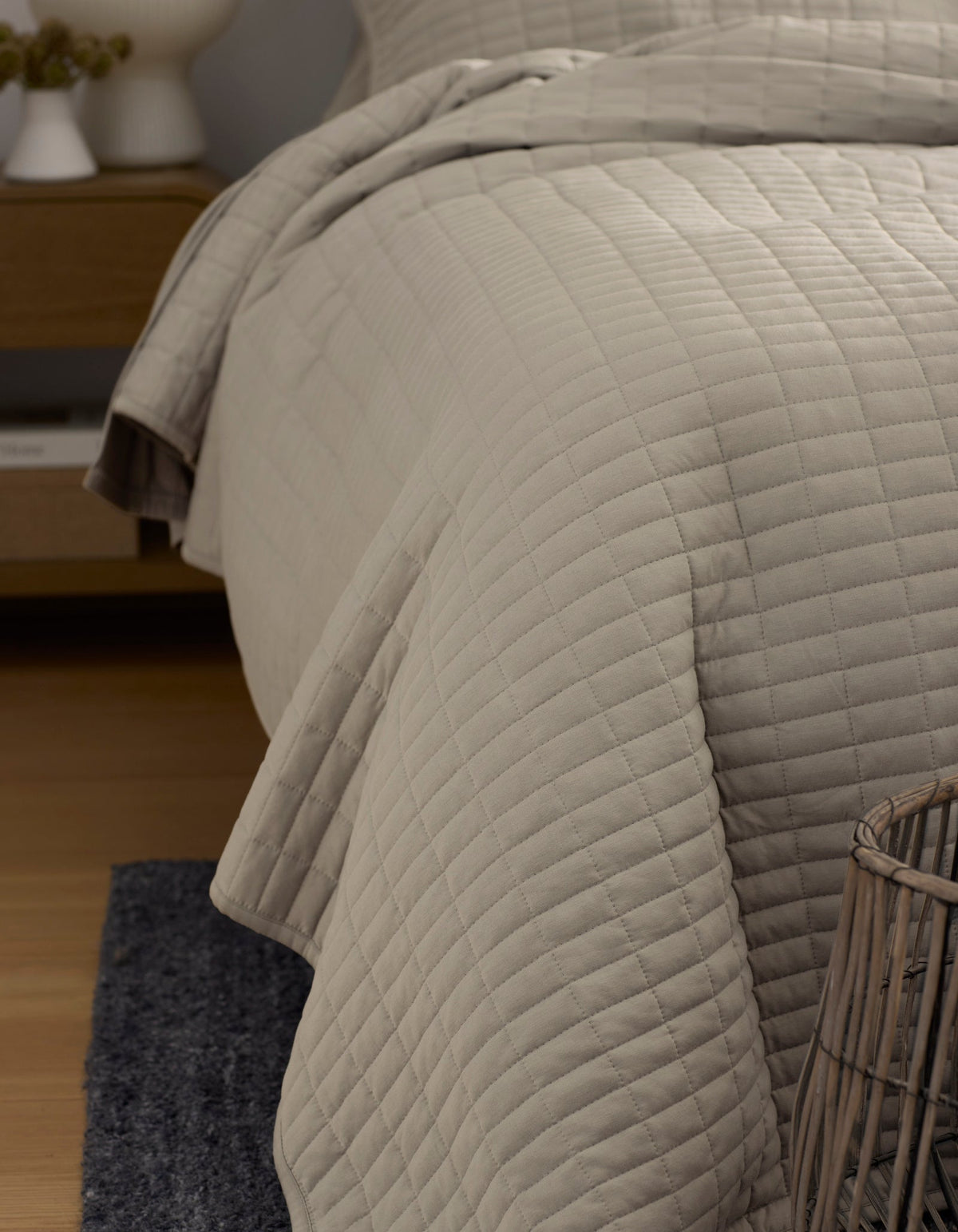 A close-up of a neatly made bed with the Cozy Earth Bamboo Jersey Quilt in light beige, featuring a square quilted pattern. A nightstand appears in the background, and a woven basket rests on a dark rug in the foreground.|Color:Driftwood