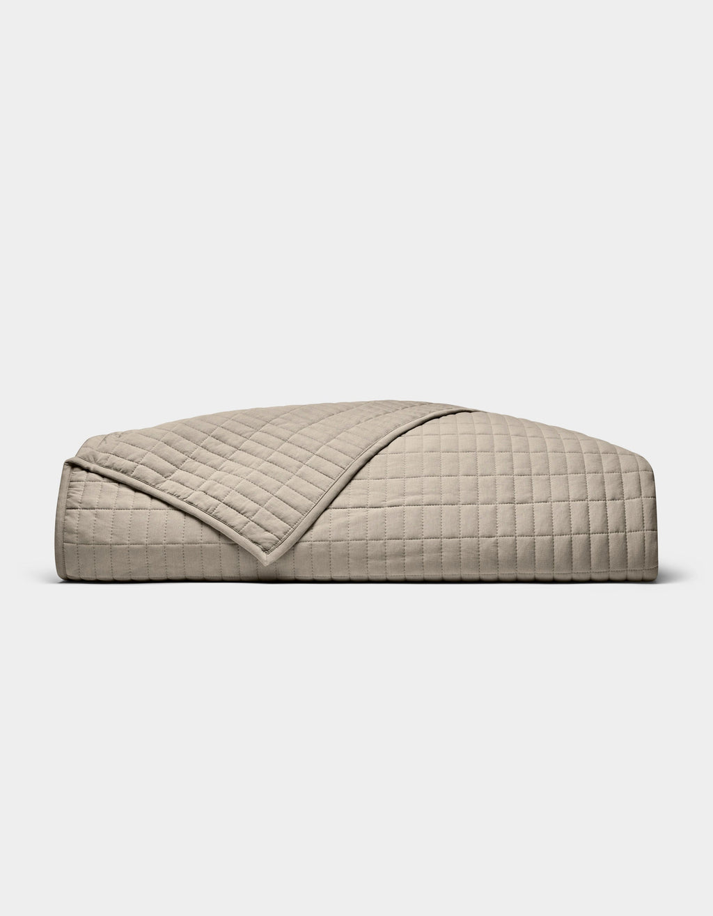 The Cozy Earth Bamboo Jersey Quilt, featuring a beige, quilted grid pattern, is neatly folded on a white background. A turned-down corner highlights its soft, luxurious texture.|Color:Driftwood