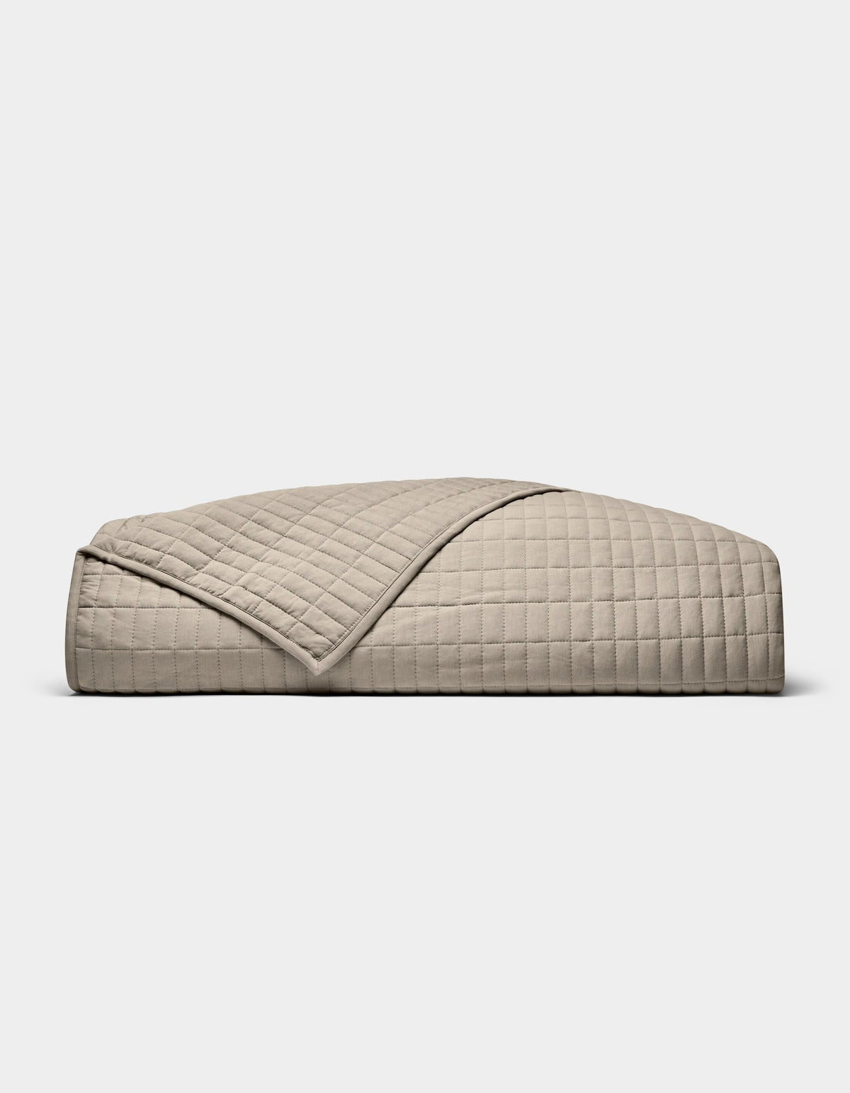 The Cozy Earth Bamboo Jersey Quilt, featuring a beige, quilted grid pattern, is neatly folded on a white background. A turned-down corner highlights its soft, luxurious texture.|Color:Driftwood