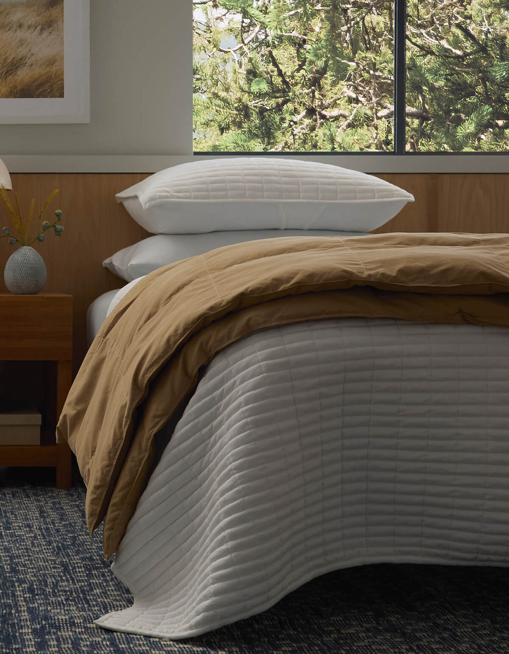 A neatly made bed features the Cozy Earth Down Alternative Throw, a tan comforter, and stacked white pillows beside a wooden nightstand. Sunlight streams through the window, revealing green trees outside.|Color:Sandalwood