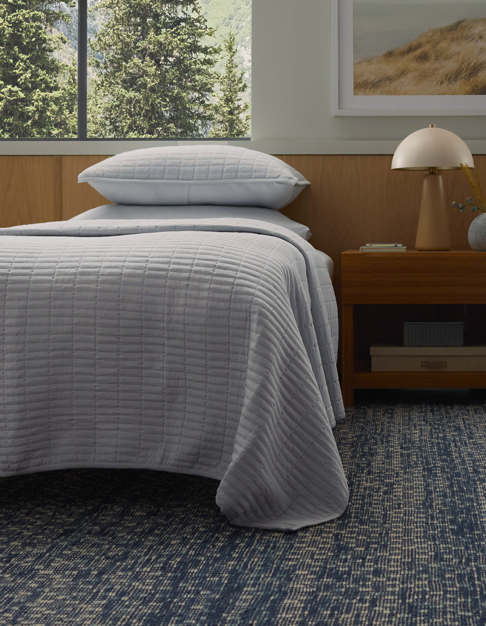 A bed with the Cozy Earth Bamboo Jersey Quilt in light gray and matching pillow is next to a wooden nightstand with a lamp, all atop a blue patterned rug. A large window and a framed landscape art piece are visible in the background.|Color:Shore