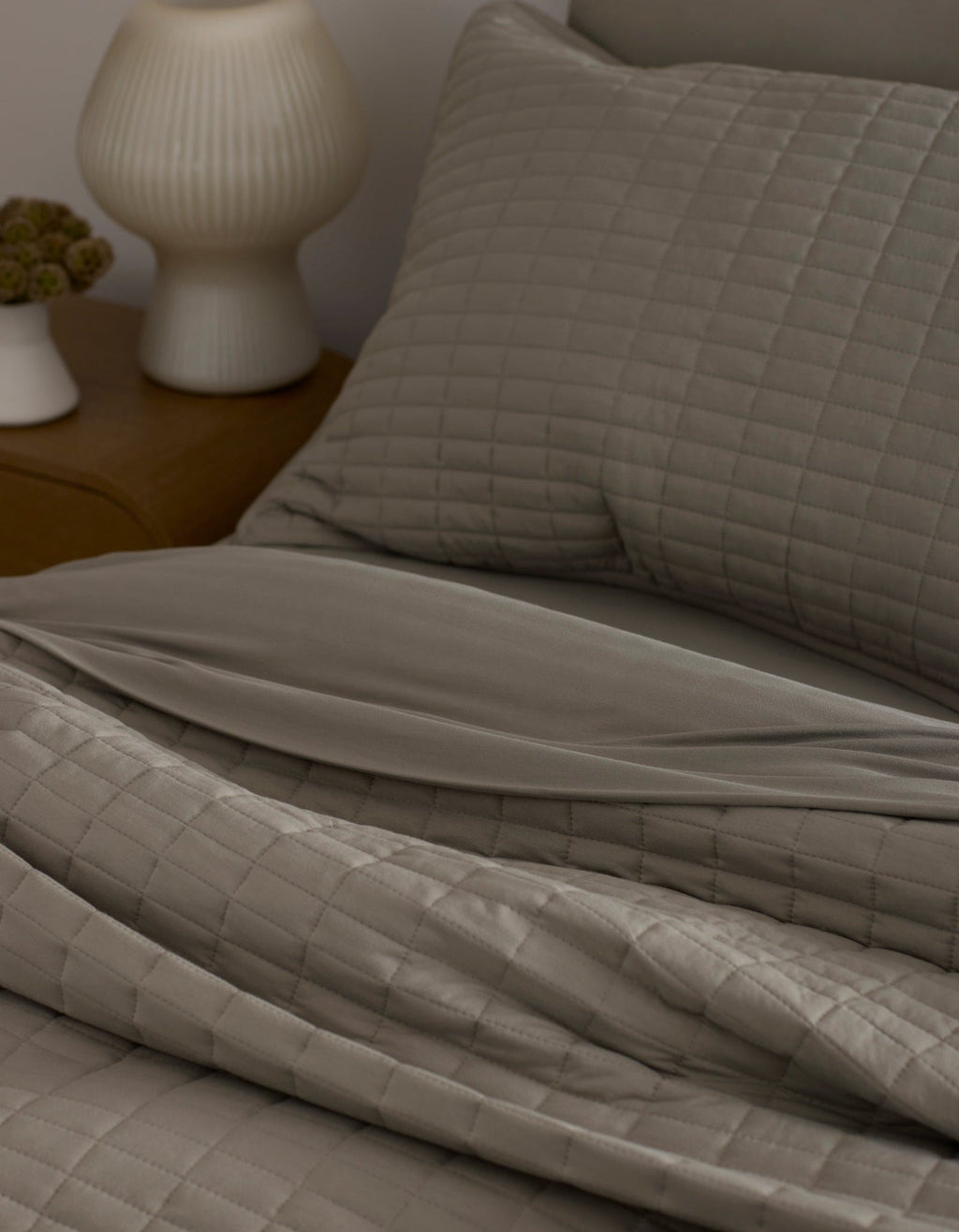 A neatly made bed features the Bamboo Jersey Quilt by Cozy Earth in taupe with matching pillows, next to a wooden nightstand topped with a white ribbed lamp and a small flower vase.|Color:Stone