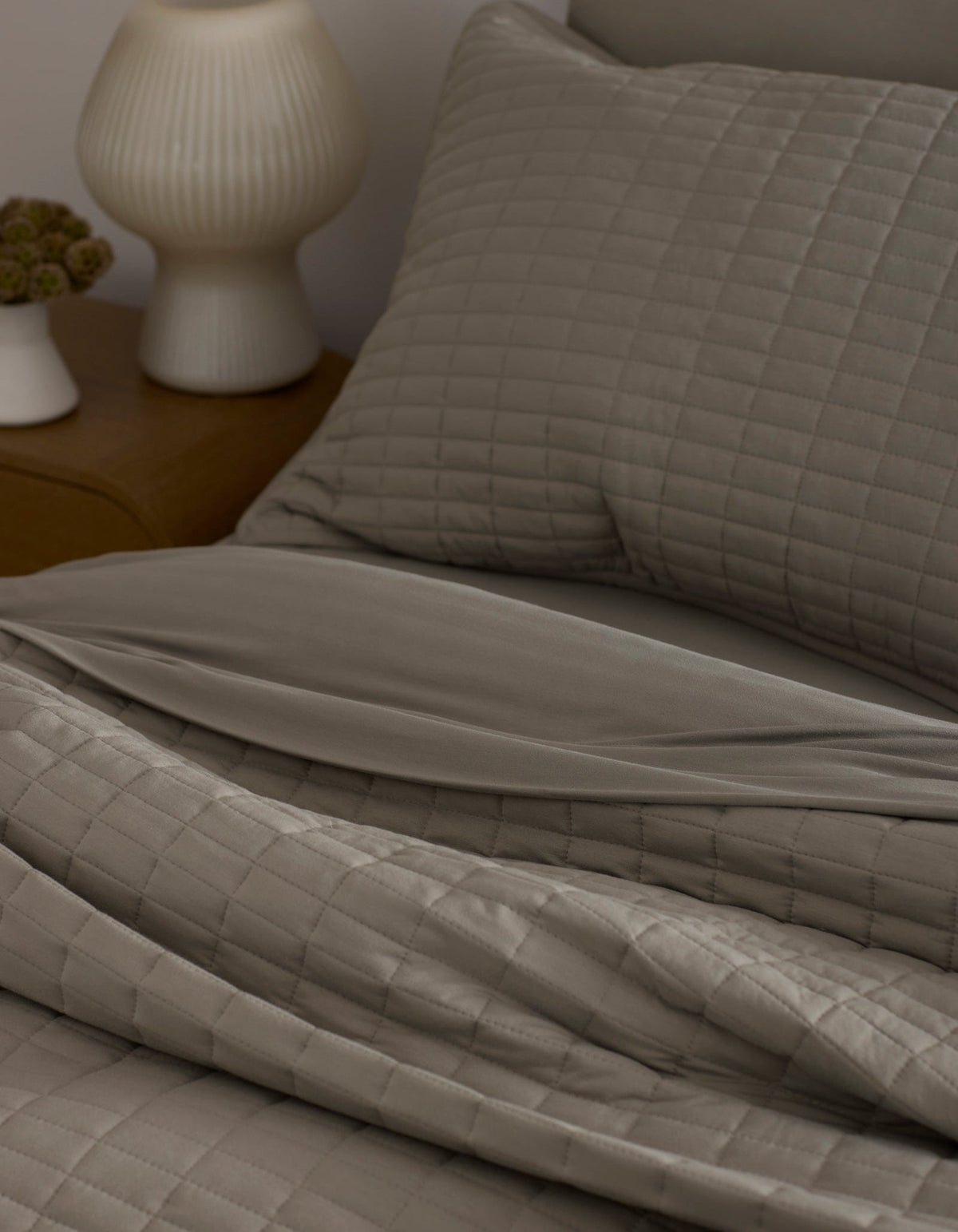 A neatly made bed features the Bamboo Jersey Quilt by Cozy Earth in taupe with matching pillows, next to a wooden nightstand topped with a white ribbed lamp and a small flower vase.|Color:Stone
