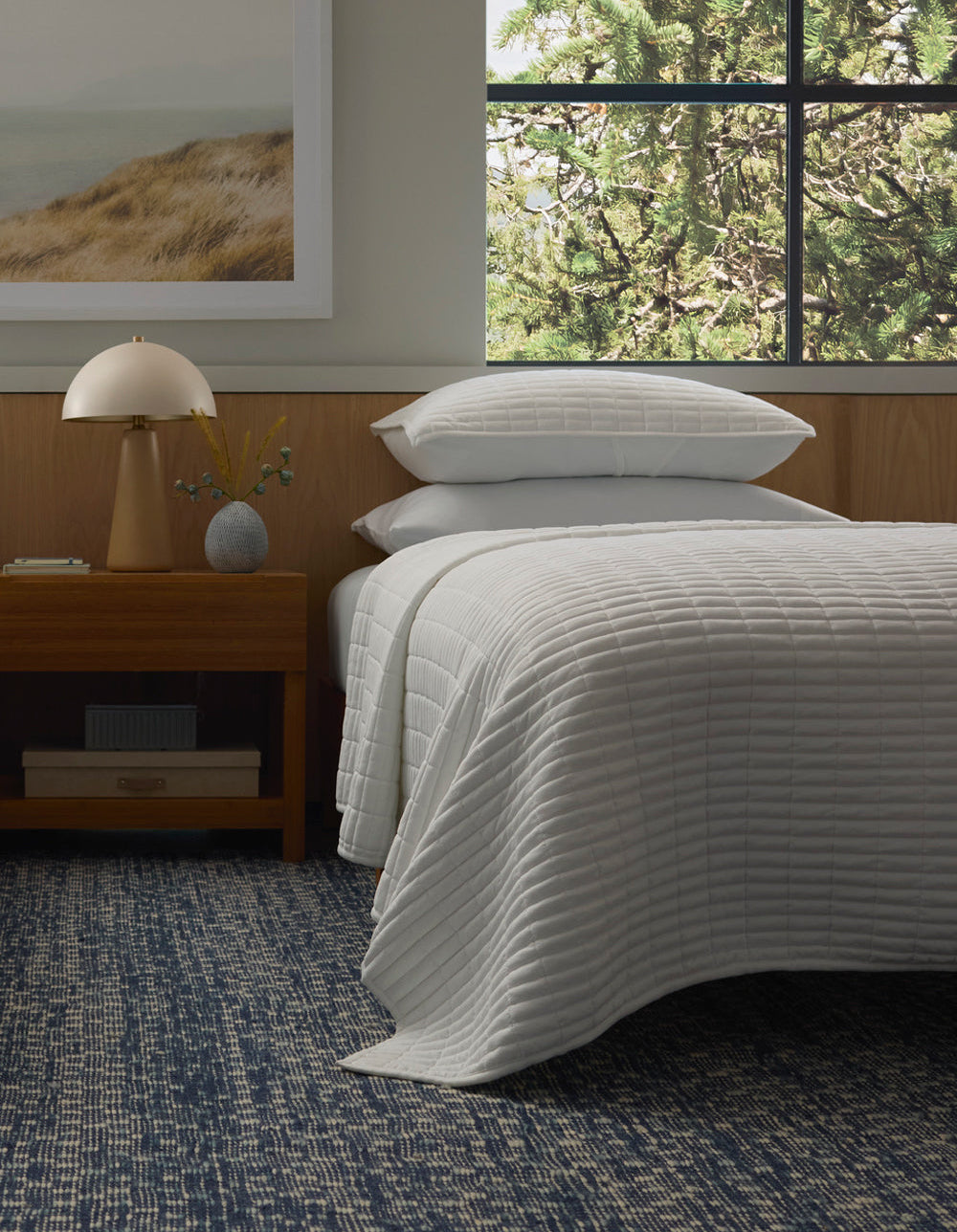 A neatly made bed features the Cozy Earth Bamboo Jersey Quilt and pillows, next to a wooden nightstand with a lamp and vase; a large window reveals green trees, while a textured blue rug covers the floor.|Color:White