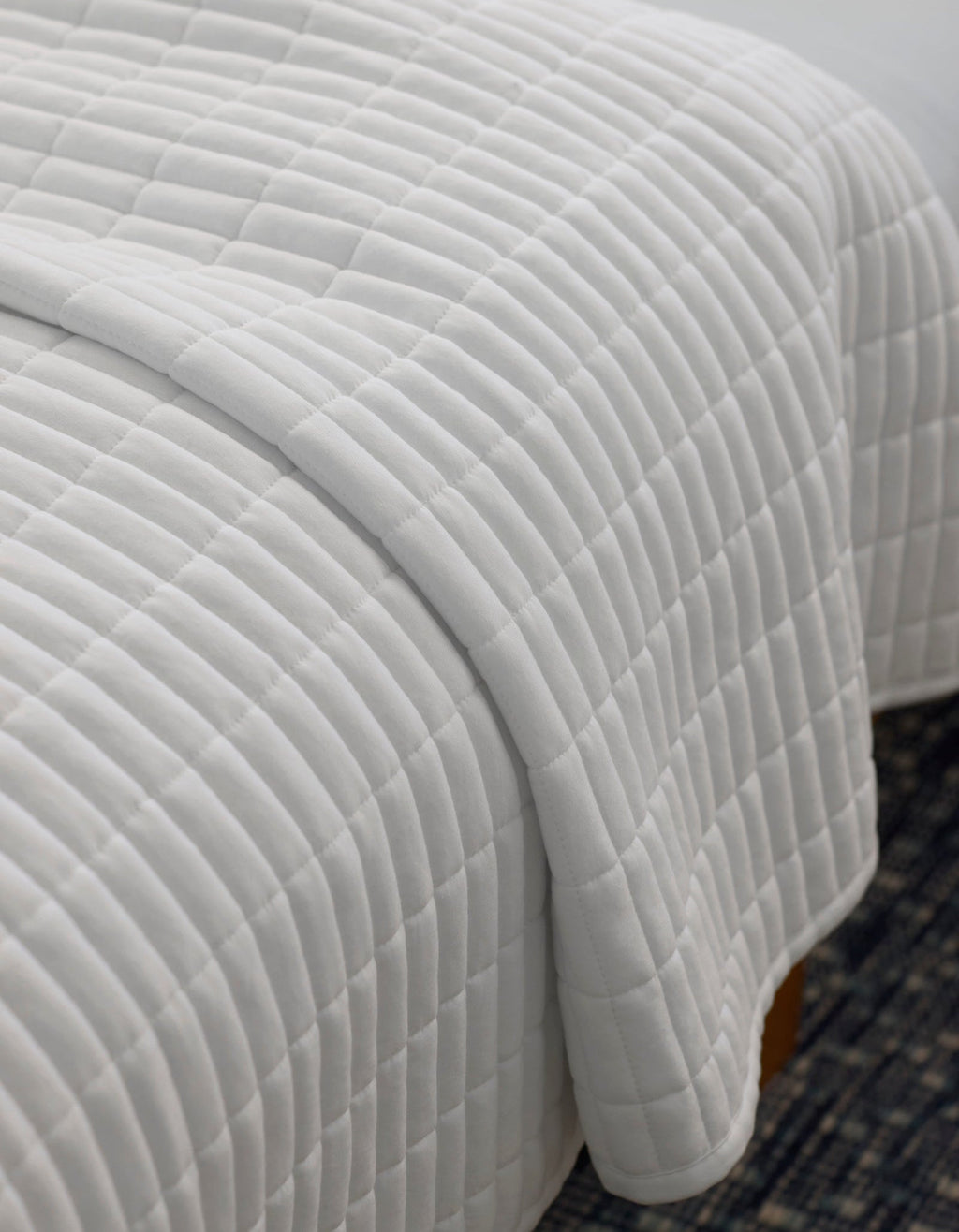 A close-up of a neatly made bed with the Cozy Earth Bamboo Jersey Quilt, featuring a white, square-patterned design, draped over the edge atop a dark patterned rug.|Color:White