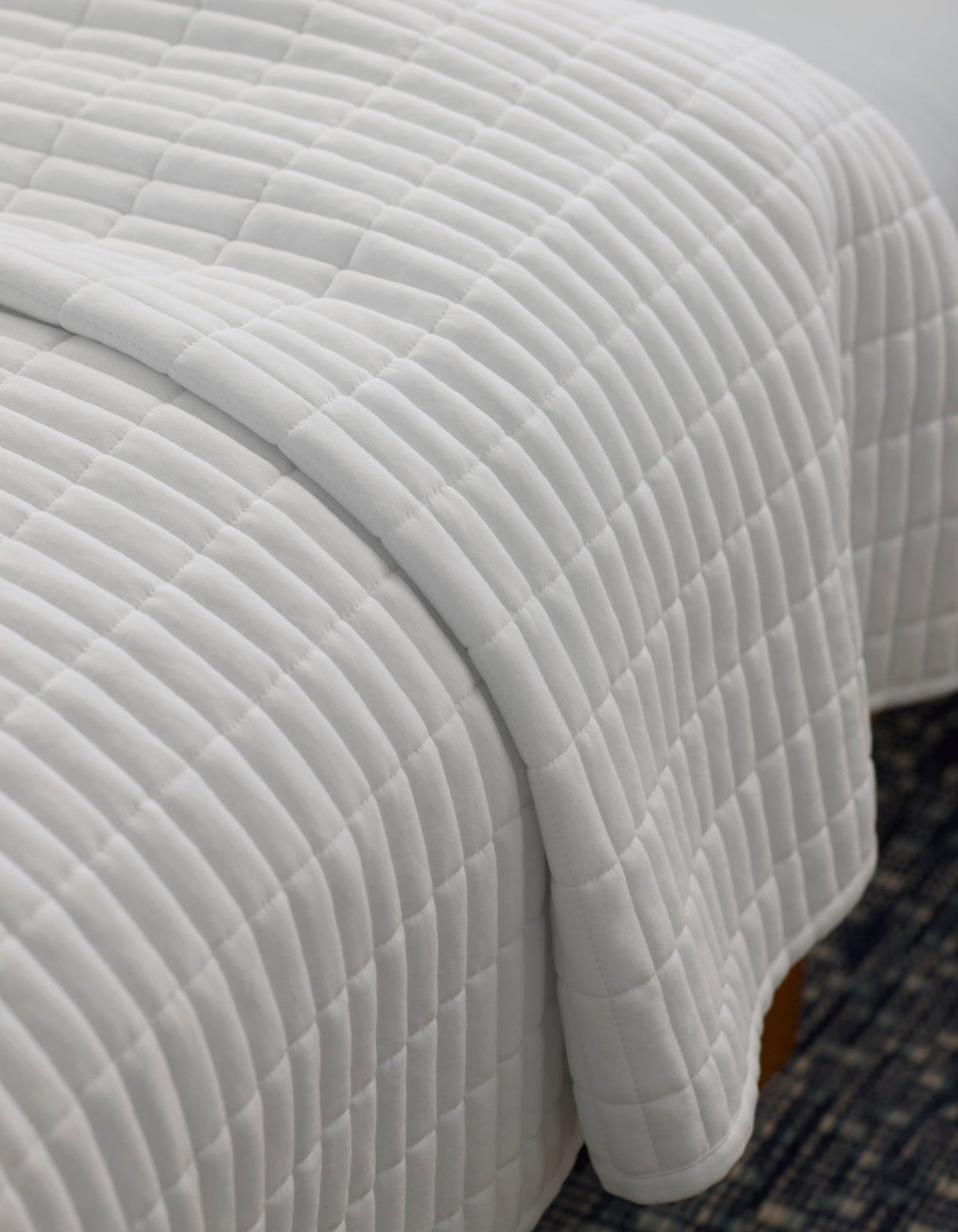 A close-up of a neatly made bed with the Cozy Earth Bamboo Jersey Quilt, featuring a white, square-patterned design, draped over the edge atop a dark patterned rug.|Color:White