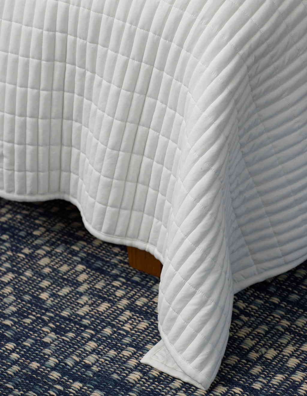 A close-up of the Cozy Earth Bamboo Jersey Quilt, featuring a white quilted grid pattern, draped over a bed with a blue and white patterned carpet underneath.|Color:White