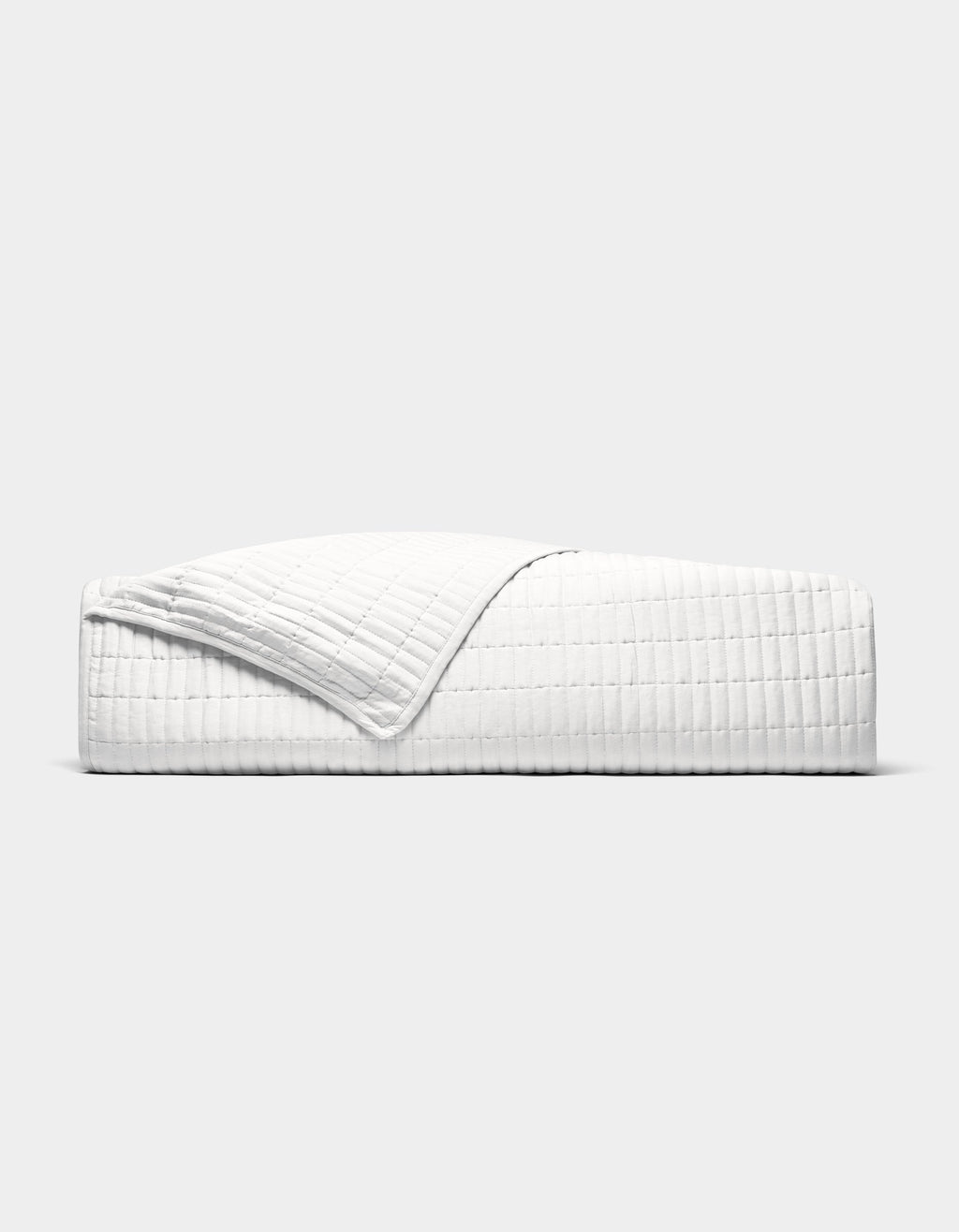 The Cozy Earth Bamboo Jersey Quilt is shown neatly made on a bed, its white, textured fabric partially folded back, photographed from the side against a plain light grey background.|Color:White