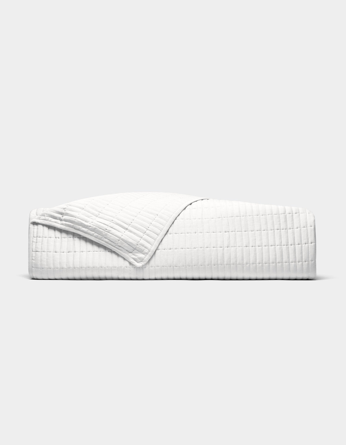 The Cozy Earth Bamboo Jersey Quilt is shown neatly made on a bed, its white, textured fabric partially folded back, photographed from the side against a plain light grey background.|Color:White