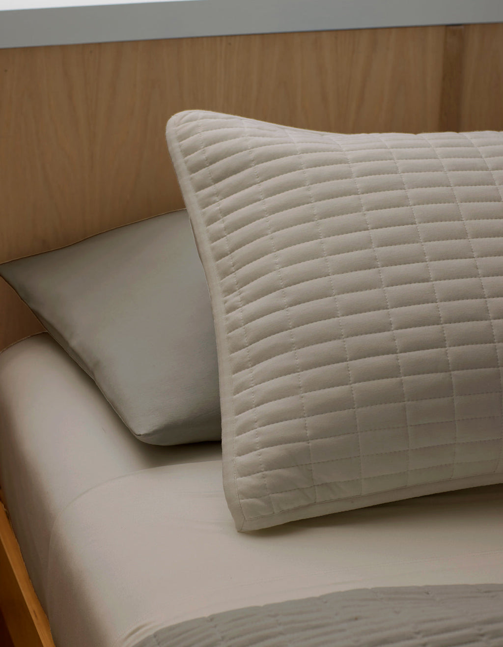 A close-up of a neatly made bed features two pillows resting against a light wooden headboard: a Bamboo Jersey Quilted Sham by Cozy Earth in beige in front, and a plain gray pillow behind.|Color:Driftwood