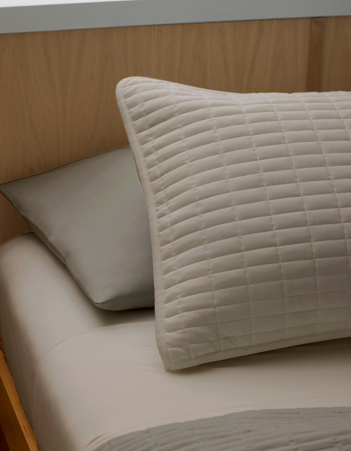 A close-up of a neatly made bed features two pillows resting against a light wooden headboard: a Bamboo Jersey Quilted Sham by Cozy Earth in beige in front, and a plain gray pillow behind.|Color:Driftwood