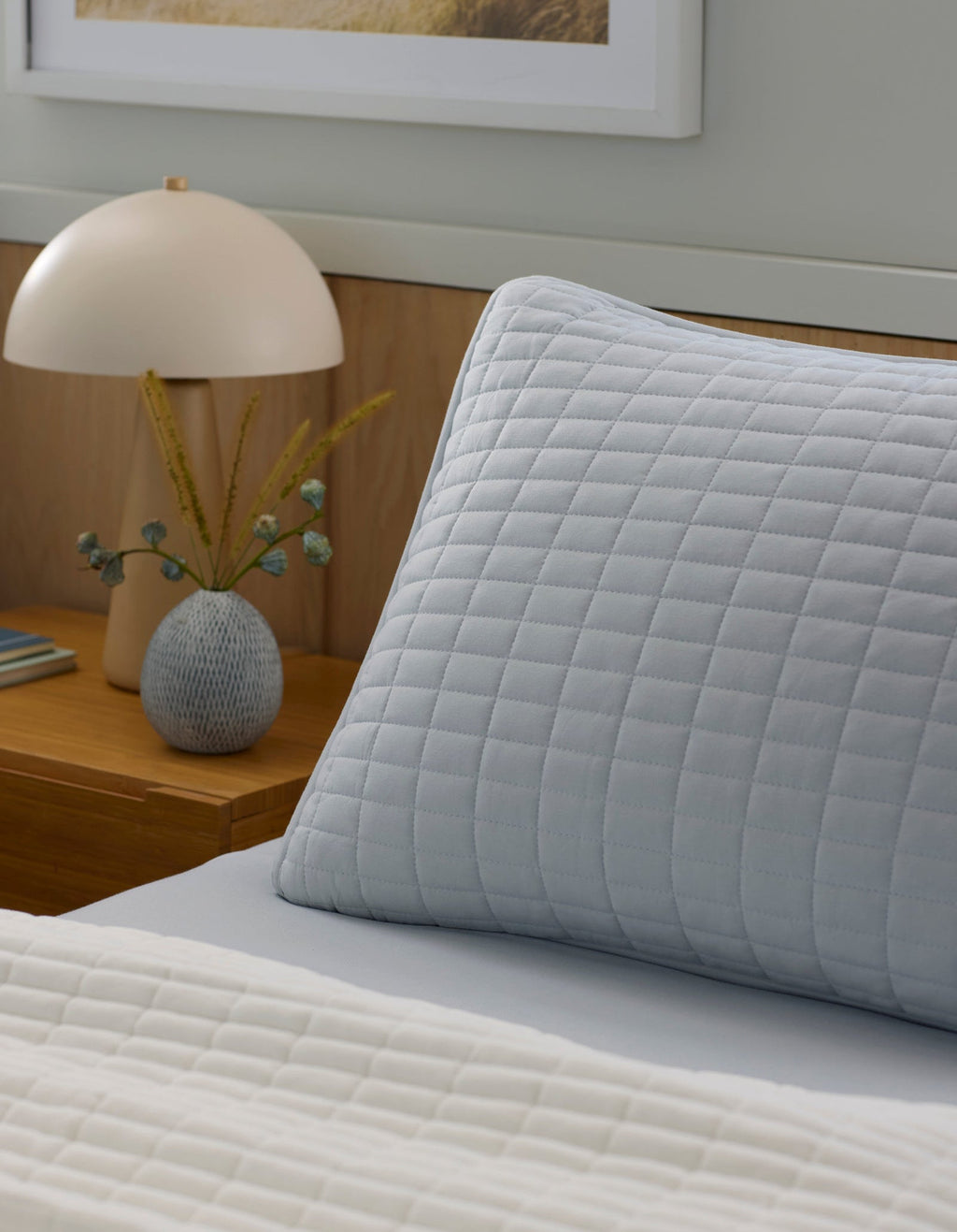 A neatly made bed features Cozy Earth Bamboo Jersey Quilted Shams in light blue and a white blanket, beside a wooden nightstand with a round lamp, small vase of dried plants, and several books.|Color:Shore