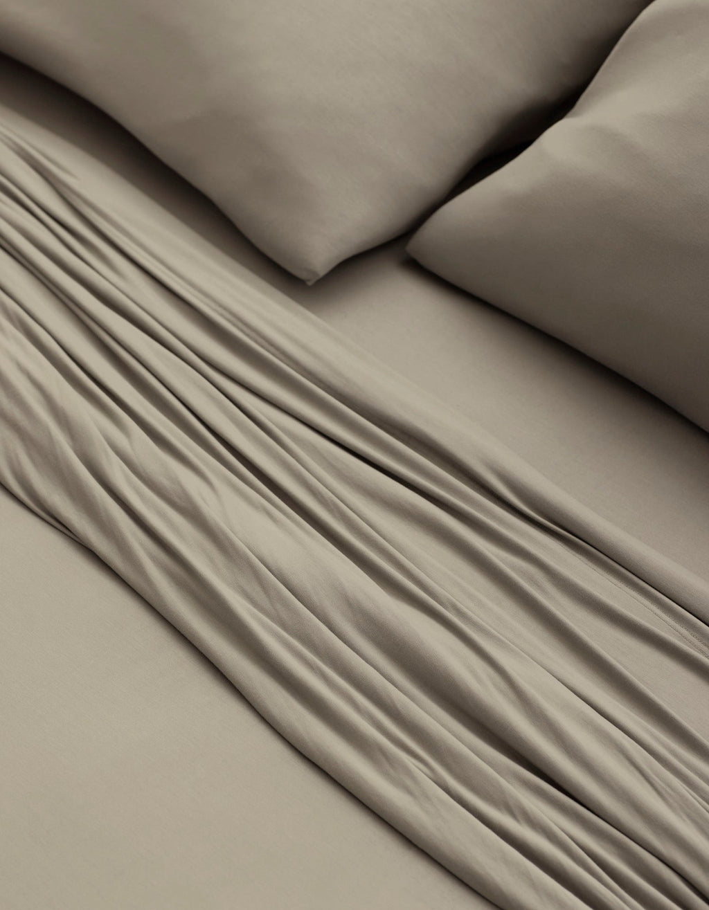 A close-up of a bed made with Cozy Earth’s Bamboo Jersey Sheet Set in taupe, featuring two matching pillows and showcasing the soft fabric folds and smooth texture.|Color:Driftwood