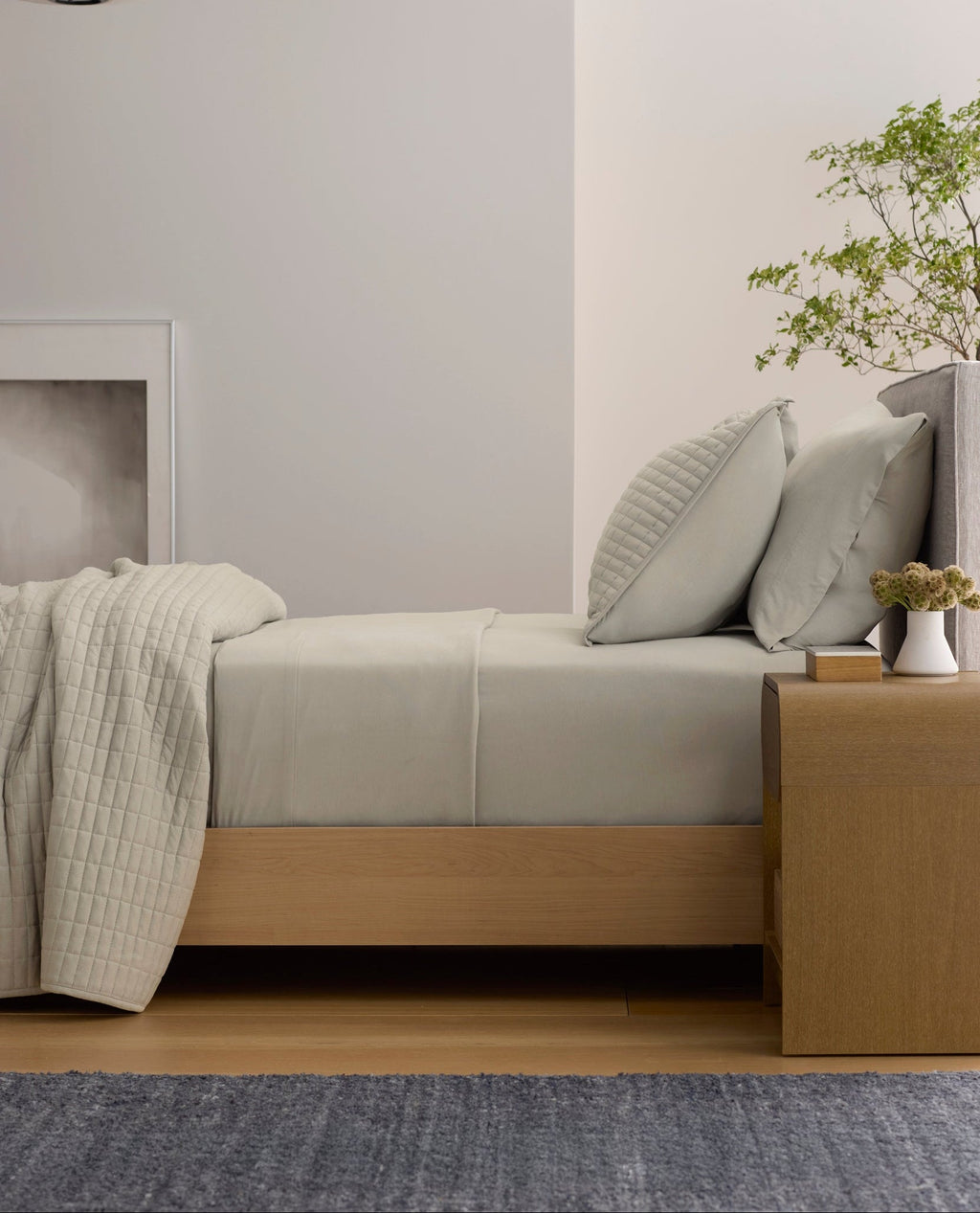 A minimalist room features a neatly made bed with Cozy Earth's Bamboo Jersey Flat Sheet in light gray, paired with pillows. Next to it is a wooden nightstand holding a small plant and vase, while a framed picture and pendant lamp complete the scene.|Color:Driftwood