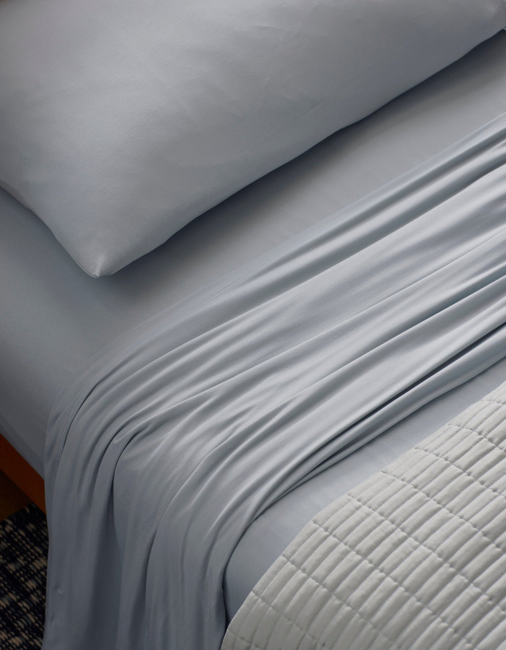 A close-up of a neatly made bed features the Cozy Earth Bamboo Jersey Sheet Set in light gray, with silky sheets and pillowcase. A textured, light quilt is folded at the foot, highlighting the smooth, soft bedding.|Color:Shore