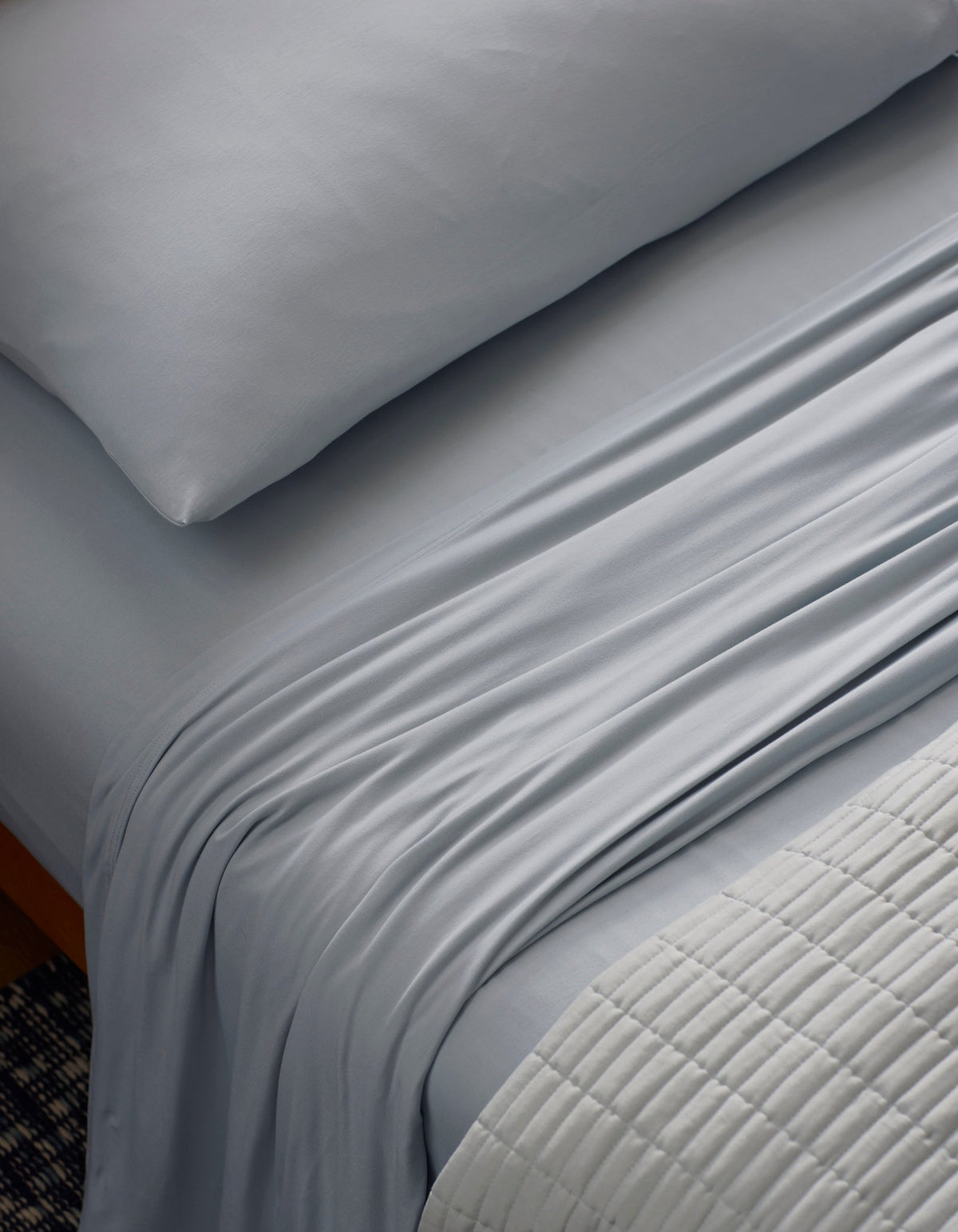 A close-up of a neatly made bed features the Cozy Earth Bamboo Jersey Sheet Set in light gray, with silky sheets and pillowcase. A textured, light quilt is folded at the foot, highlighting the smooth, soft bedding.|Color:Shore