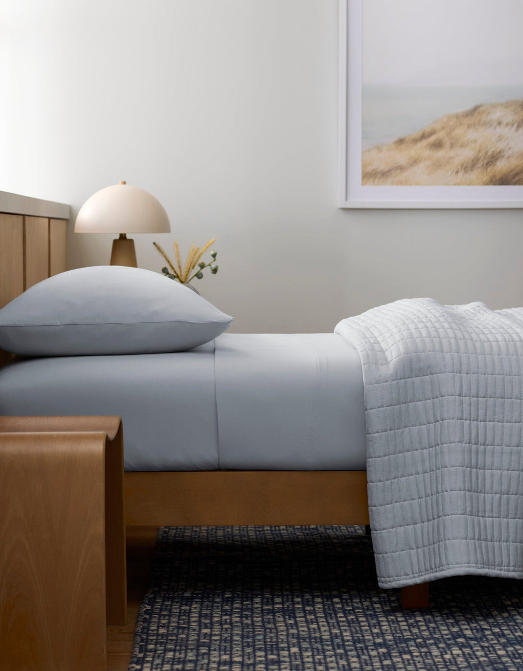 A minimalist bedroom features a neatly made bed with Cozy Earth's Bamboo Jersey Flat Sheet in light gray, a wooden bench, a small lamp, and a landscape dunes photo; natural light enhances the serene, neutral space.|Color:Shore