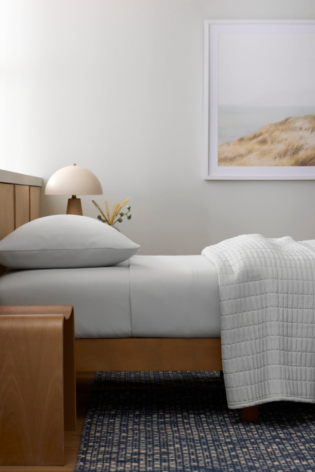 A modern, minimal bedroom features the Cozy Earth Bamboo Jersey Fitted Sheet in light gray, a wooden bench, a small round table lamp, a framed beach landscape on the wall, and a blue patterned rug on the floor.|Color:White