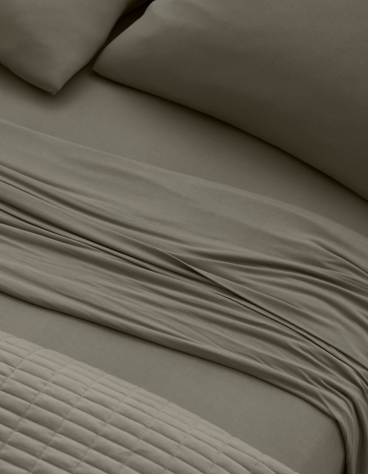 Close-up of a neatly made bed with Cozy Earth Bamboo Jersey Sheet Set in taupe, featuring pillows, a slightly rumpled flat sheet, and a textured quilted blanket at the foot of the bed.|Color:Stone