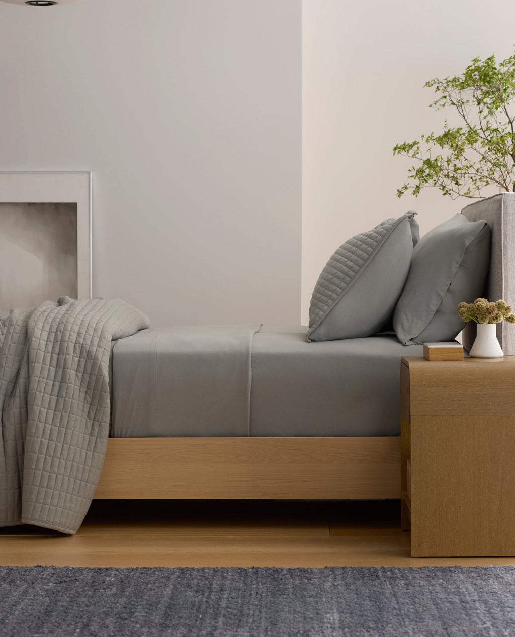 A neatly made bed features the Cozy Earth Bamboo Jersey Flat Sheet in gray, paired with pillows. Next to it, a wooden nightstand holds a potted plant, books, and a vase, while a hanging light and minimalist decor create a calm, modern bedroom.|Color:Stone