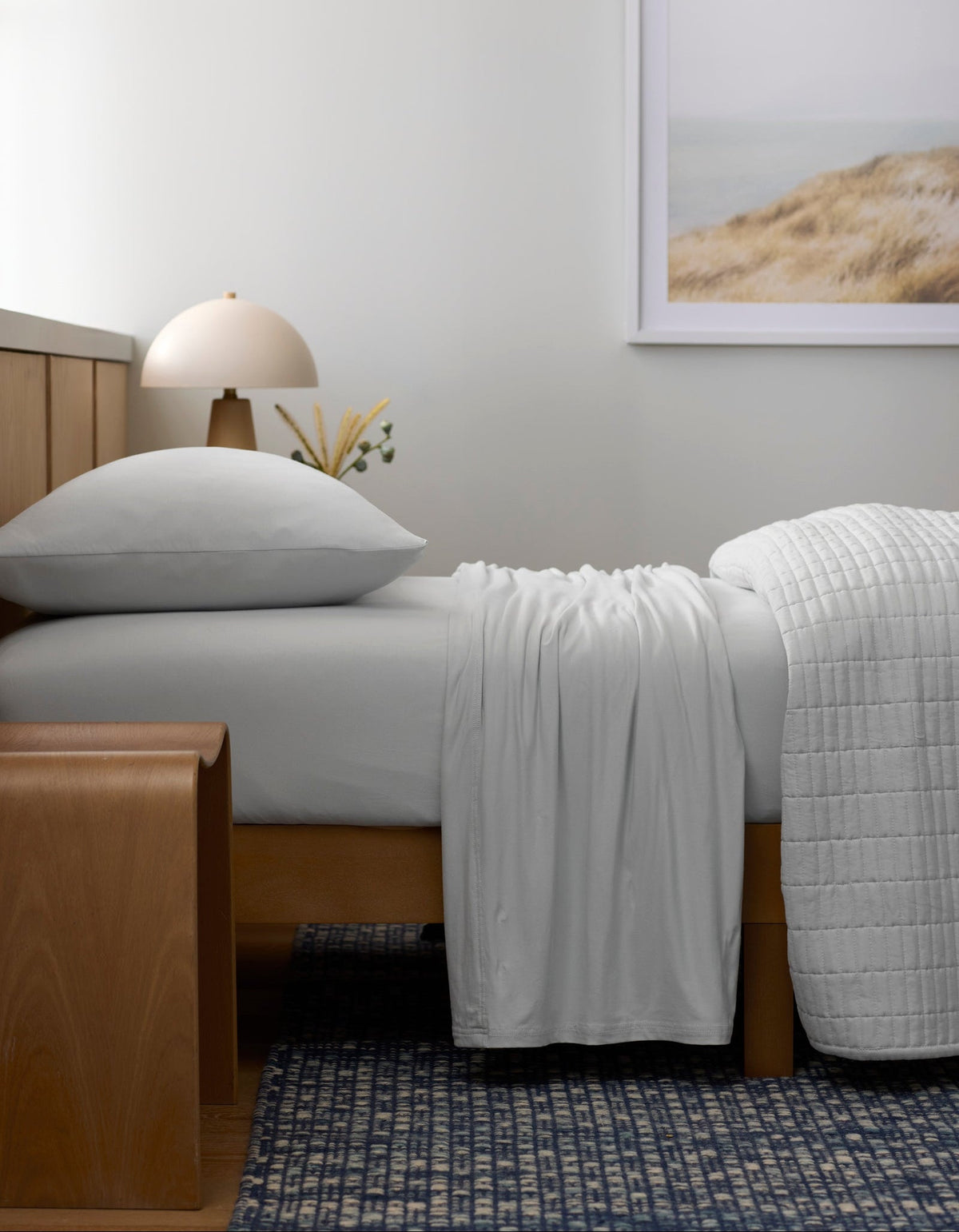 A bed made with Cozy Earth’s Bamboo Jersey Sheet Set rests against a wooden headboard. A wooden bench stands at the foot, while a lamp, small vase, and framed landscape photo accentuate the minimal, serene bedroom.|Color:White