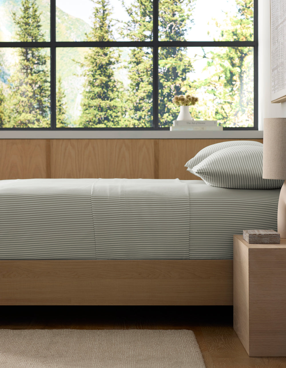 A neatly made bed featuring Cozy Earth’s Bamboo Fitted Sheet sits beside a wooden nightstand and lamp, with large windows framing views of tall evergreen trees and mountains in the background.|Color:Classic Stripe in Laurel