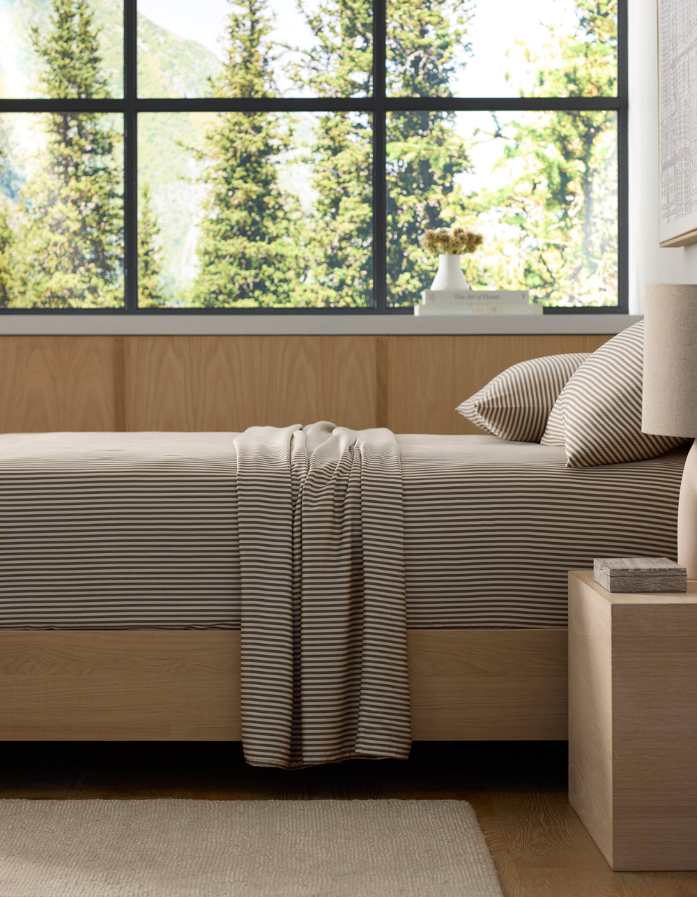 A neatly made bed features Cozy Earth's Bamboo Sheet Set in beige and white stripes, set in a modern bedroom with light wood furniture. Large windows showcase green trees and mountains, filling the room with natural light.|Color:Classic Stripe in Ebony