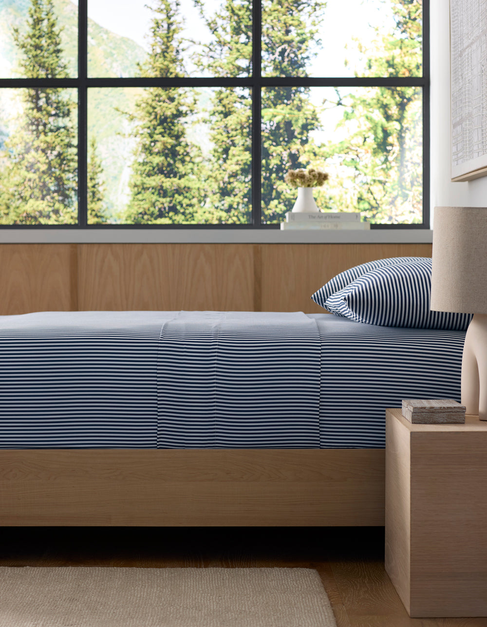 A neatly made bed with Cozy Earth's Bamboo Fitted Sheet is next to a wooden nightstand holding a lamp and book. Large windows offer views of green trees and mountains outside.|Color:Classic Stripe in Pacific