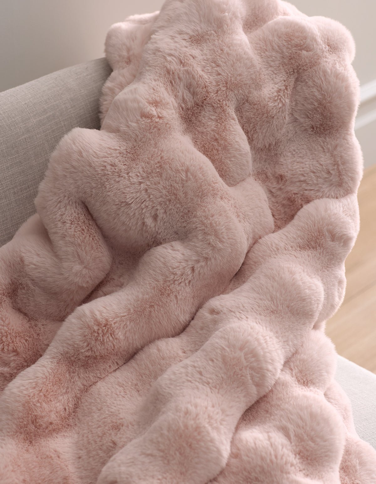 The Bubble Cuddle Blanket by Cozy Earth, featuring soft, plush light pink faux fur with a wavy texture, is draped over the arm of a light gray upholstered chair.|Color:Blossom