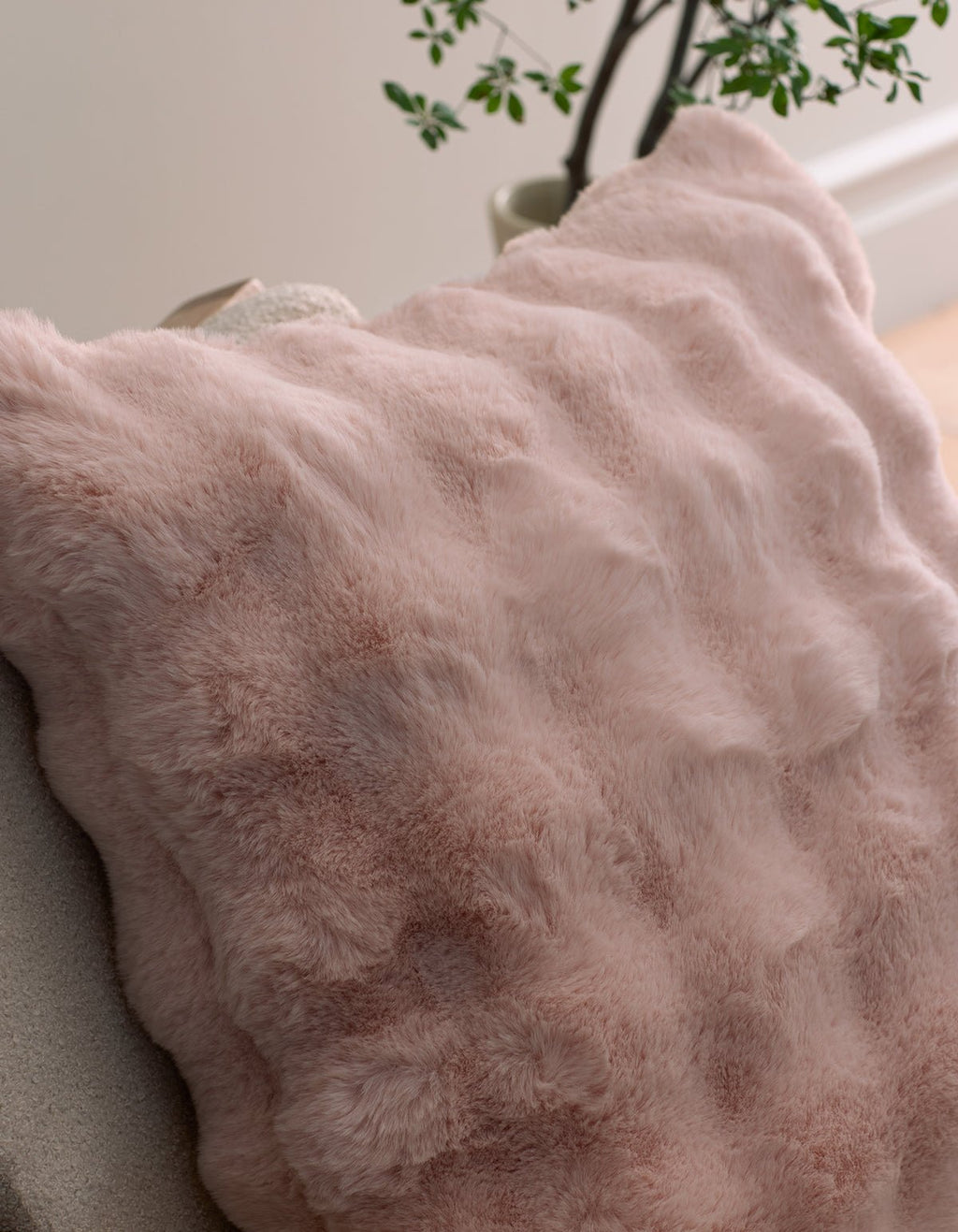 A close-up of the Bubble Cuddle Pillow by Cozy Earth in soft light pink faux fur rests on a chair, with a leafy plant and a pale wall in the background.|Color:Blossom |Size:Euro