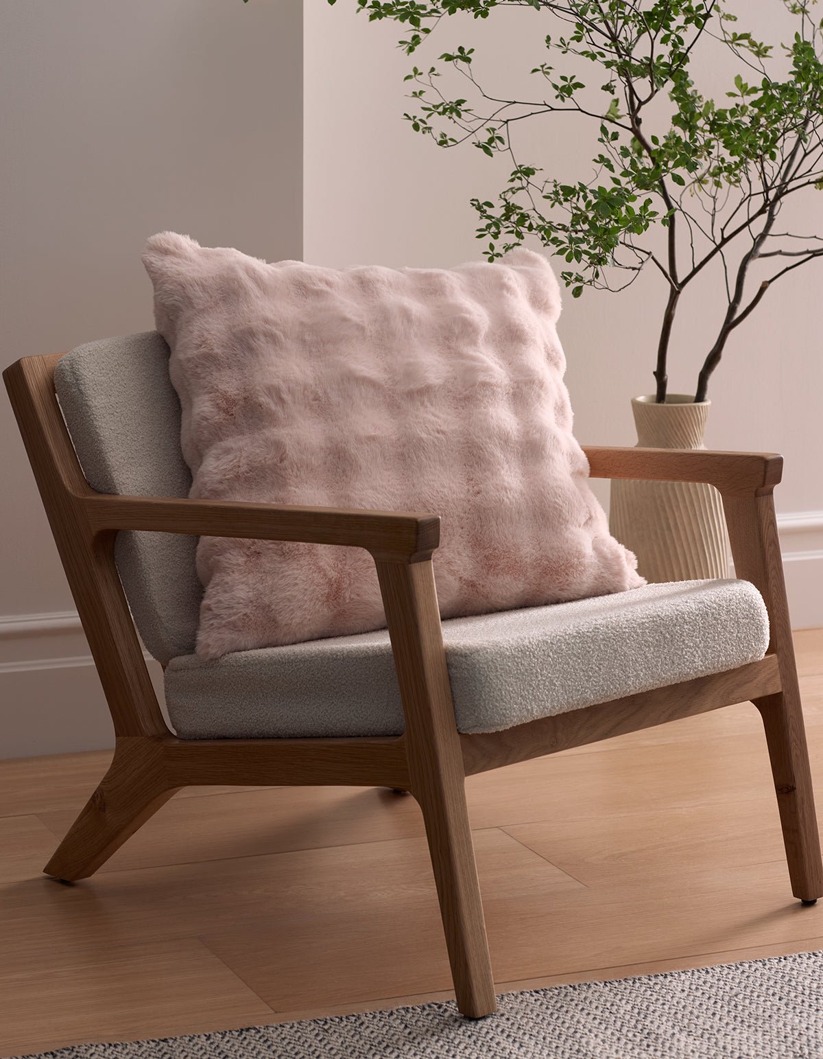 A modern wooden armchair with light gray upholstery sits on a wood floor, featuring the Bubble Cuddle Pillow by Cozy Earth. A leafy green plant in a cream vase stands beside the chair against a pale wall.|Color:Blossom |Size:24" x 24"