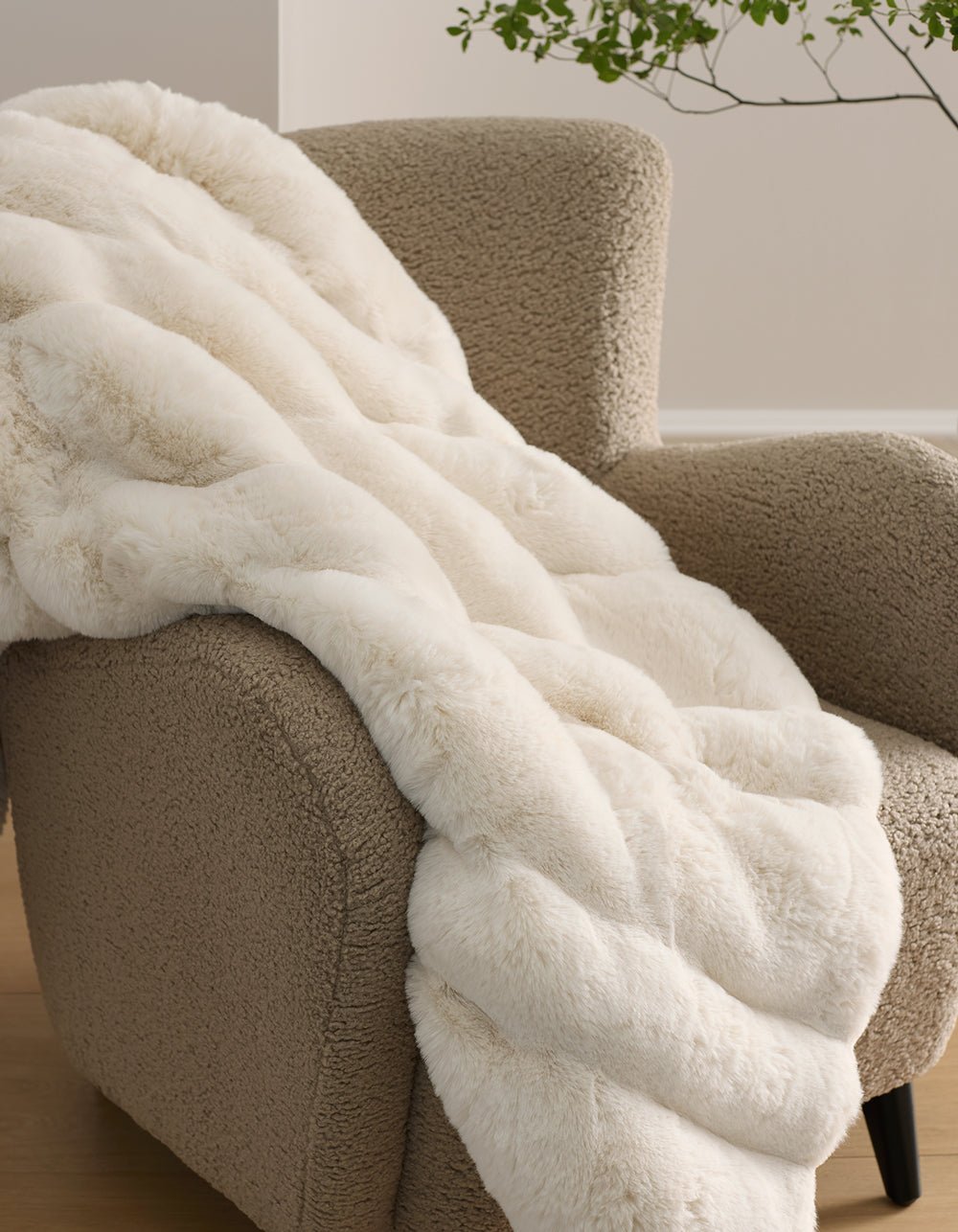 The Cozy Earth Channel Cuddle Blanket, a plush cream-colored faux fur throw, is draped over a textured beige armchair in a cozy modern room with a plant in the background.|Color:Creme