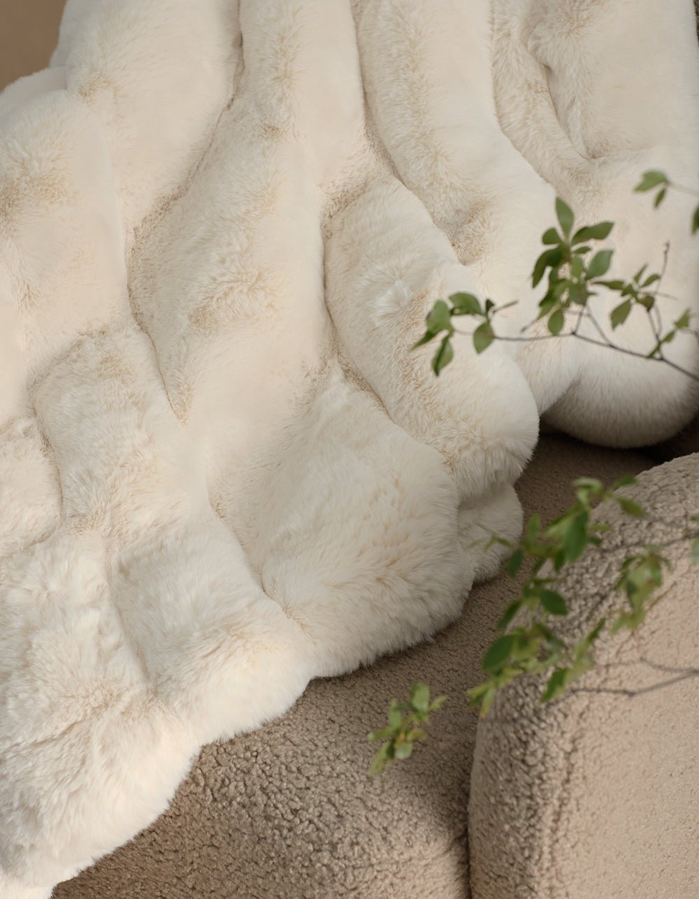The Cozy Earth Channel Cuddle Blanket, in soft creamy white faux fur, is draped over a textured beige chair with a green leafy plant in the foreground.|Color:Creme